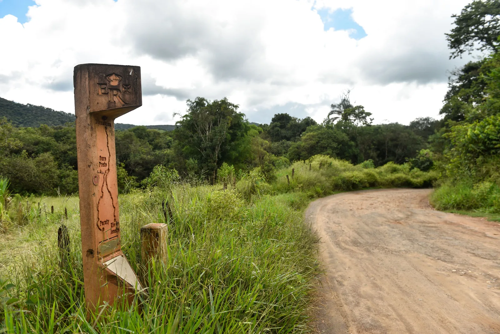 A carved wooden waymarker post standing in long grass beside a red dirt track curving away through green scrub and low trees on the Estrada Real near Ouro Preto