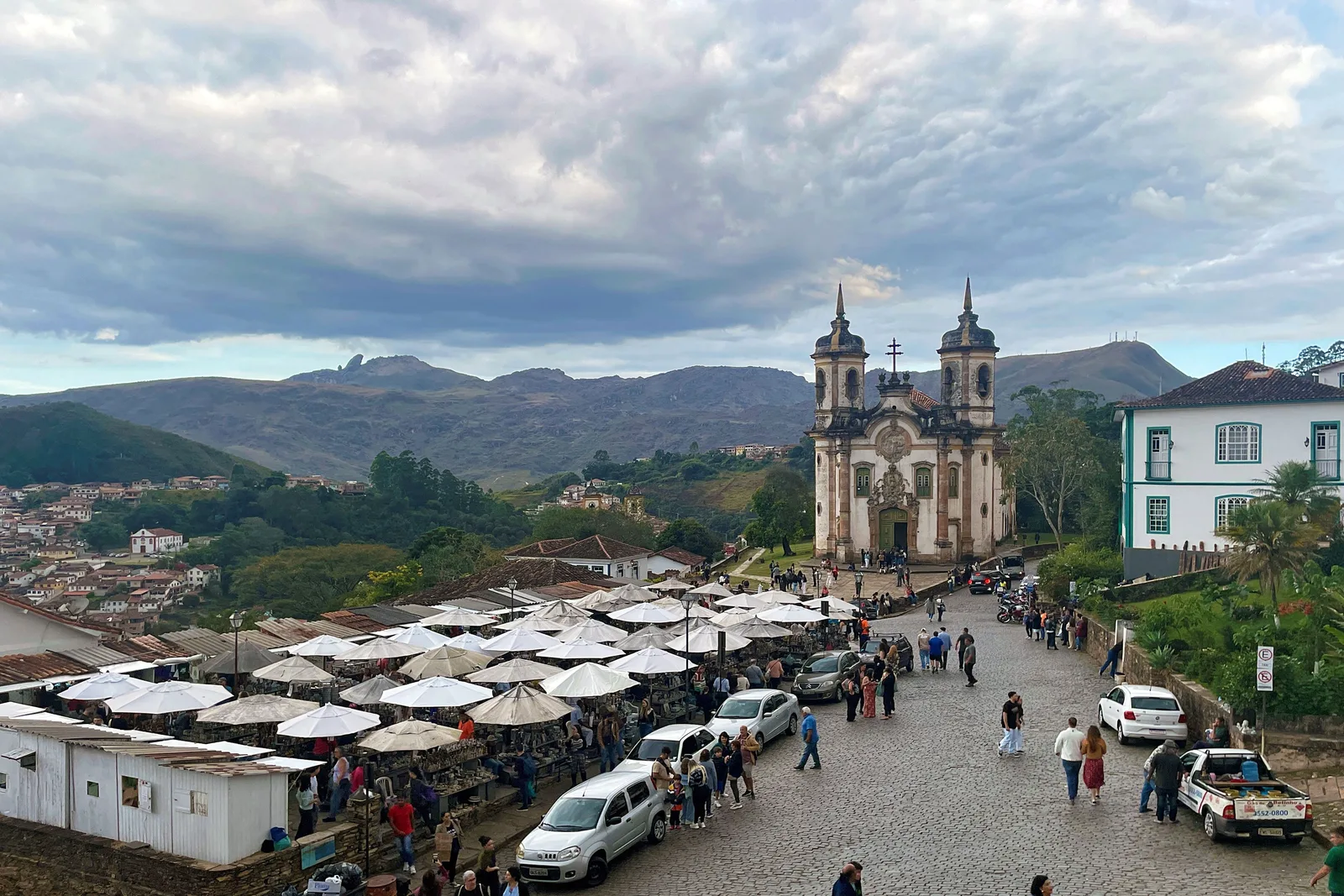 The soapstone market under rows of white umbrellas on the open square below the Igreja de São Francisco de Assis in Ouro Preto, the church standing beyond the stalls with the Pico do Itacolomi on the skyline behind it