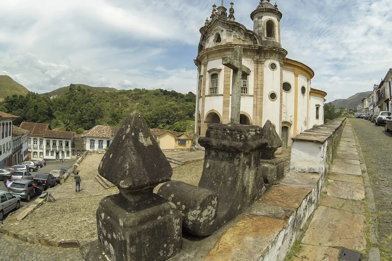 The Igreja de Nossa Senhora do Rosário in Ouro Preto seen from below on its slope: a curved white baroque facade between two rounded bell towers, a dark carved stone monument in the foreground and green hills rising behind the rooftops