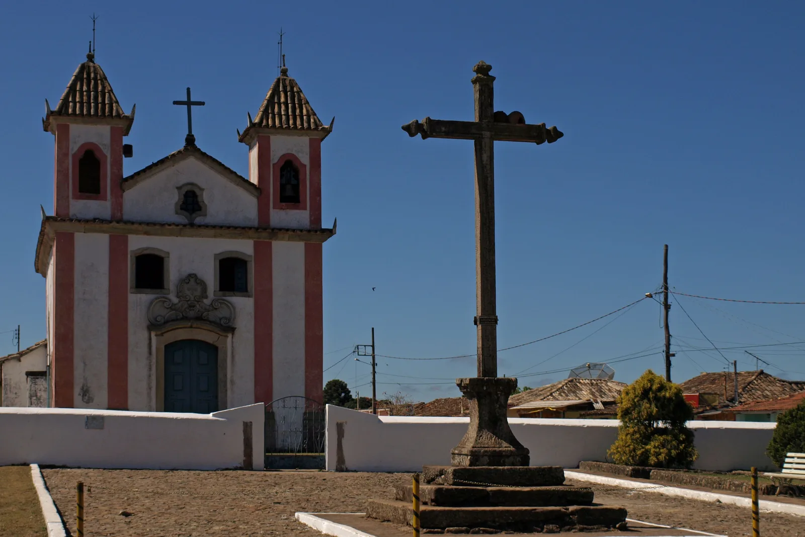 The small white church at Lavras Novas with two low bell towers, a tall dark wooden cross standing on a stone base in the open square in front of it against a deep blue sky