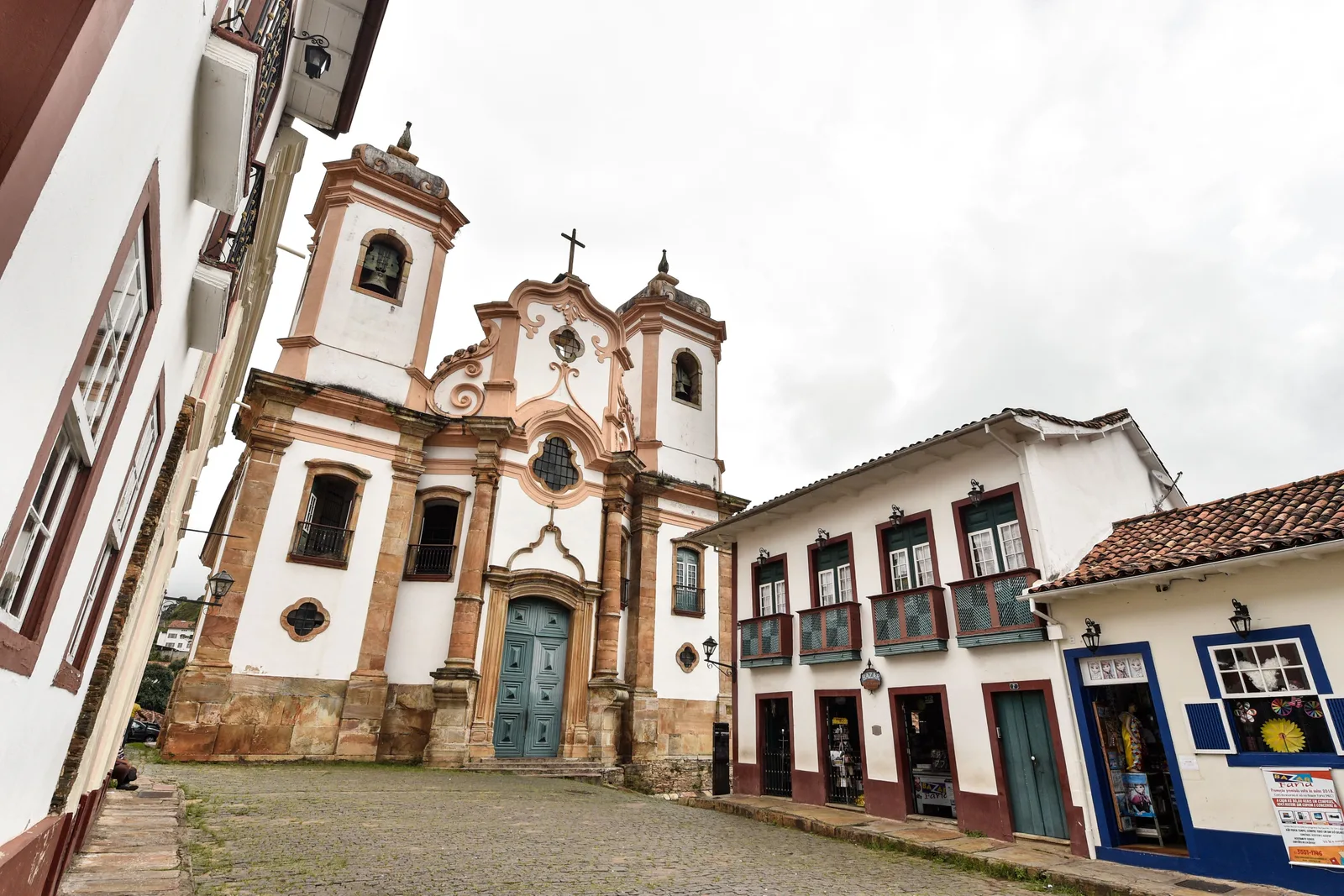 The white twin-towered facade of the Matriz de Nossa Senhora do Pilar standing at the head of a steep cobbled street in Ouro Preto, colonial shopfronts and painted houses running down either side toward the camera