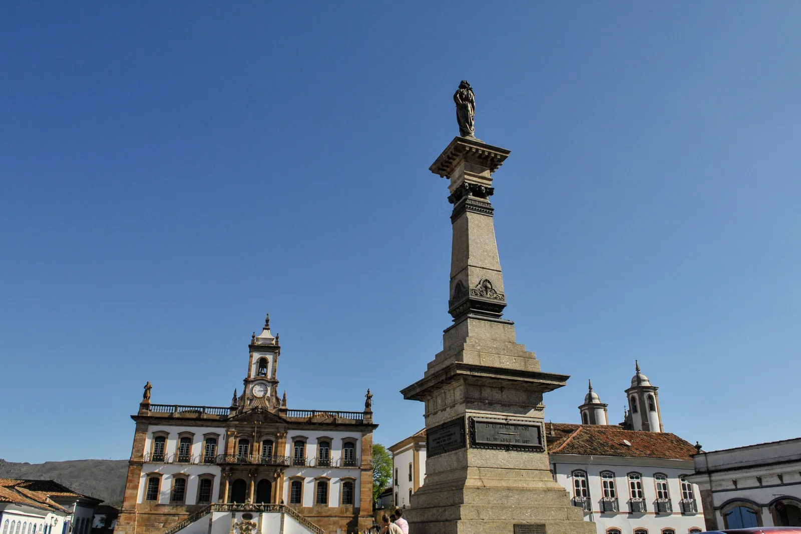 The tall stone Tiradentes column standing in the middle of Praça Tiradentes in Ouro Preto against a clear sky, the long facade of the Museu da Inconfidência behind it and colonial houses and a church tower along the far side of the square