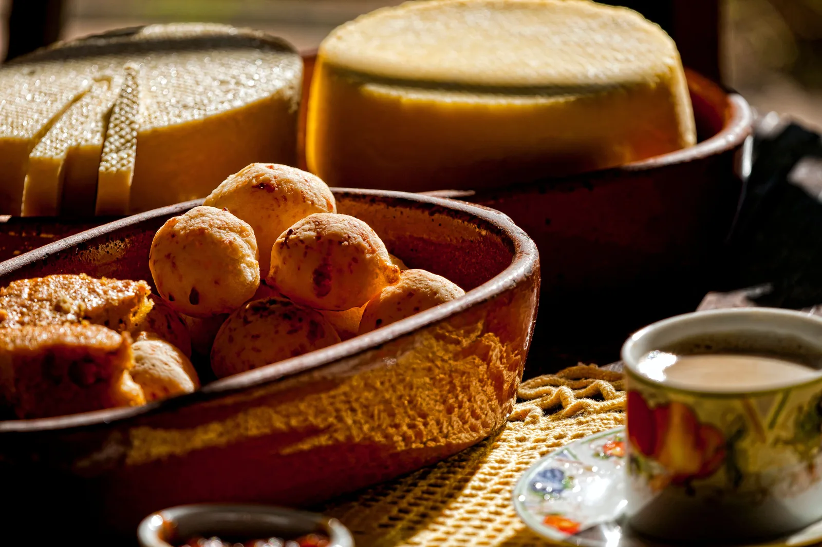 A wooden bowl of golden pão de queijo on a breakfast table beside a cup of black coffee, a slice of cake and a pale wheel of Minas cheese behind it in warm low light
