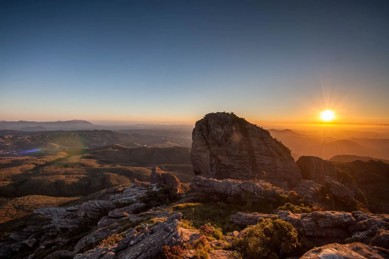 Sunrise from the rocks at the top of the Pico do Itacolomi, bare boulders in the foreground and the sun breaking over layered blue ridges and a valley of low mist below