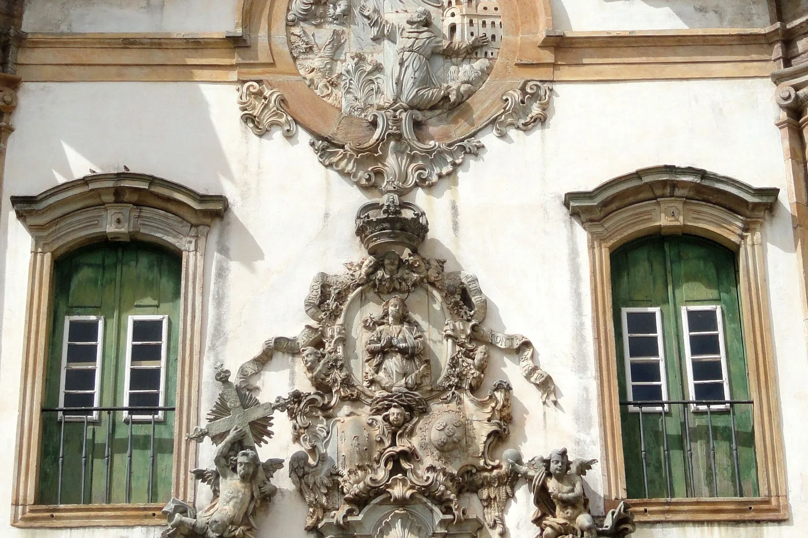 Aleijadinho’s carved soapstone medallion on the facade of São Francisco de Assis, a deeply cut relief of figures and scrollwork under a crown, set into whitewashed stone between two tall windows with green shutters