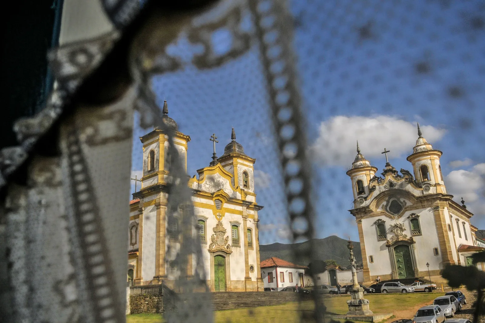 The two baroque churches of Mariana’s Praça Minas Gerais seen through an out-of-focus wrought-iron screen, their pale twin-towered facades standing at an angle to each other across the paved square