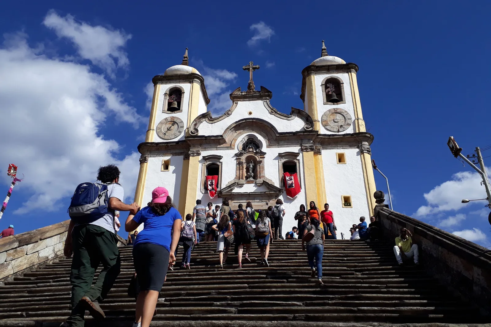 Visitors climbing the wide stone stairway to the Igreja de Santa Efigênia in Ouro Preto, a white church with two bell towers and a clock face standing above them against a bright sky with scattered cloud