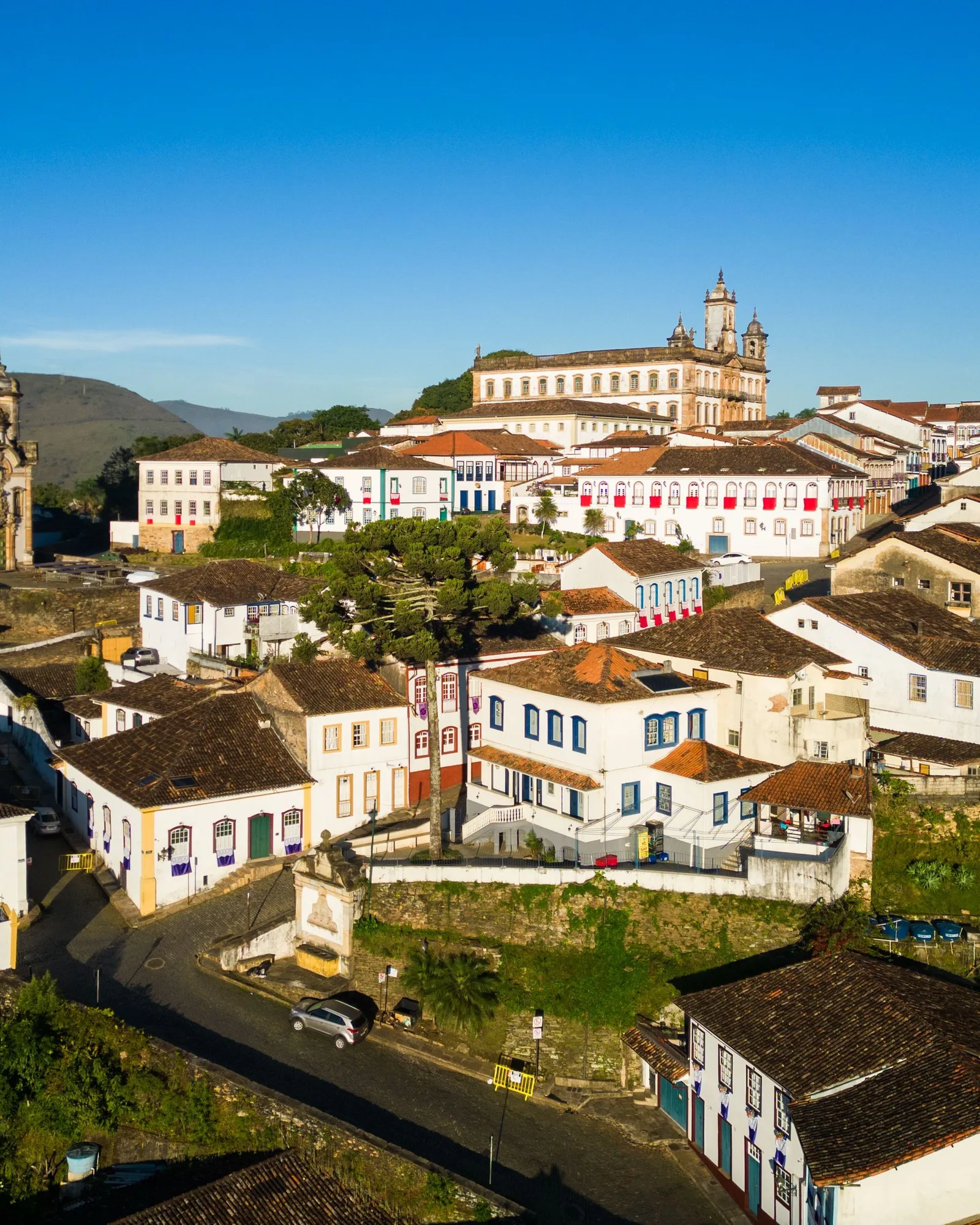 Ouro Preto in morning light: white colonial houses with red-tiled roofs stacked up the hillside below the long facade of the Museu da Inconfidência and a baroque church tower on the ridge, under a clear blue sky