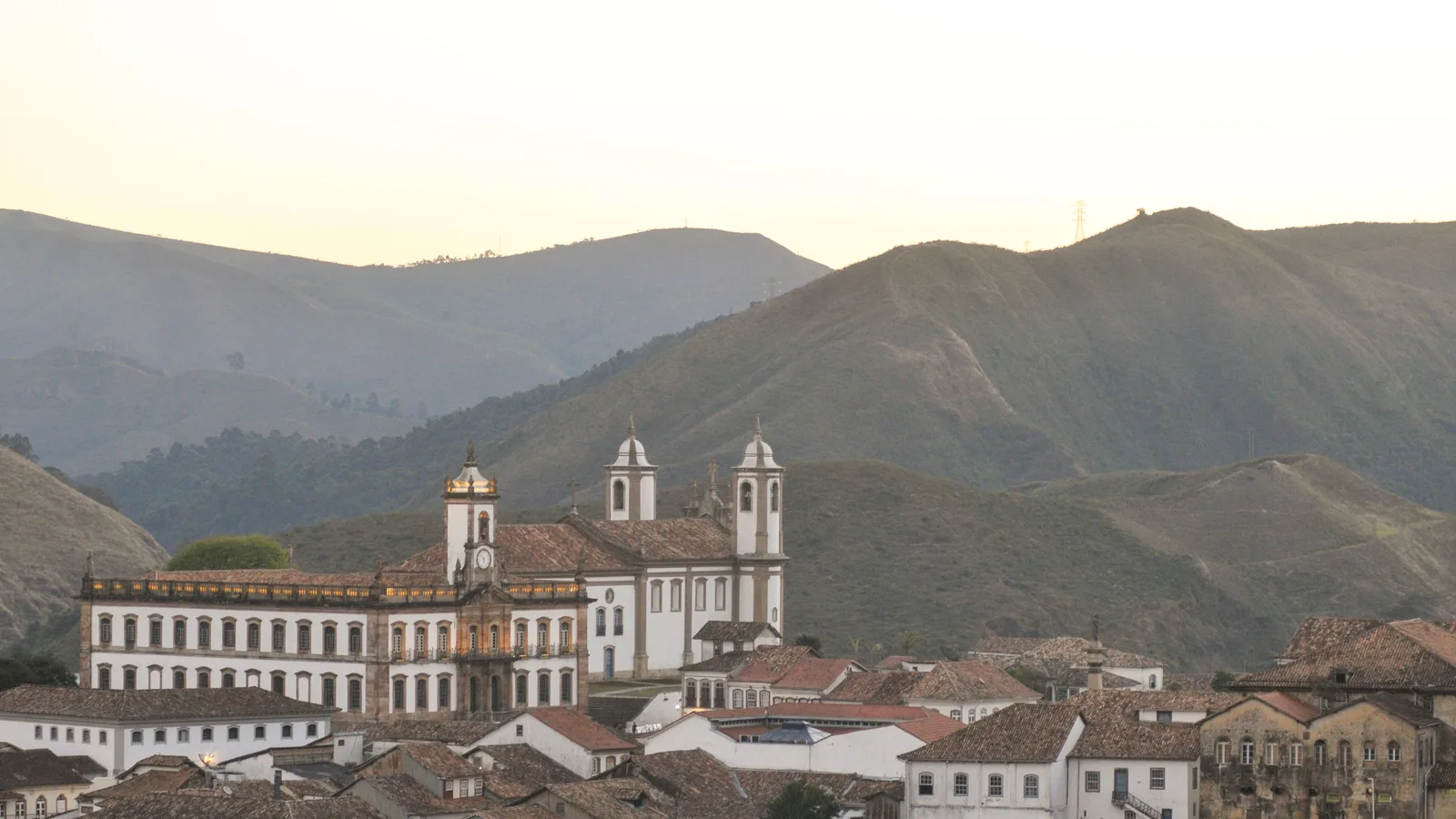 Ouro Preto in hazy late light seen across the valley: white colonial houses and red roofs stacked along the slope with a twin-towered baroque church standing among them, and layered blue mountains filling the background