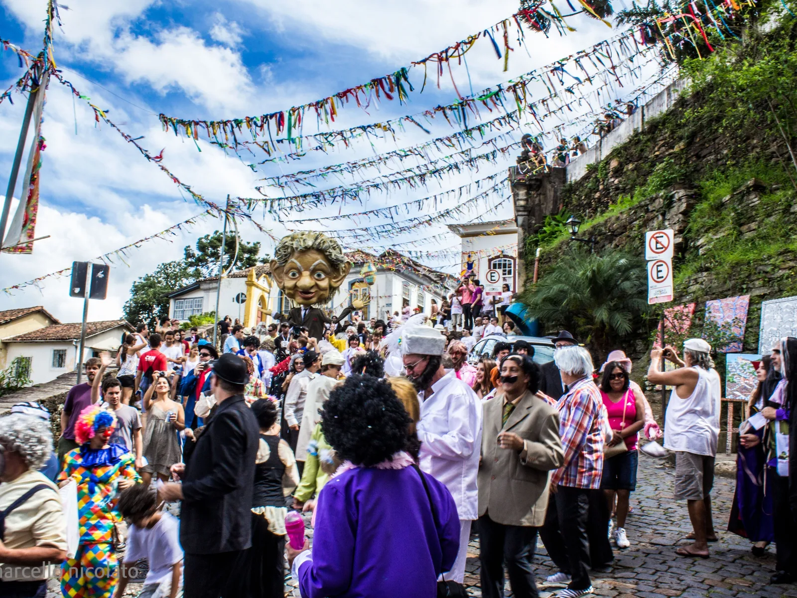 A Carnival crowd in costume filling a cobbled Ouro Preto street under strings of bunting, colonial houses on both sides and a green hillside of houses rising behind them