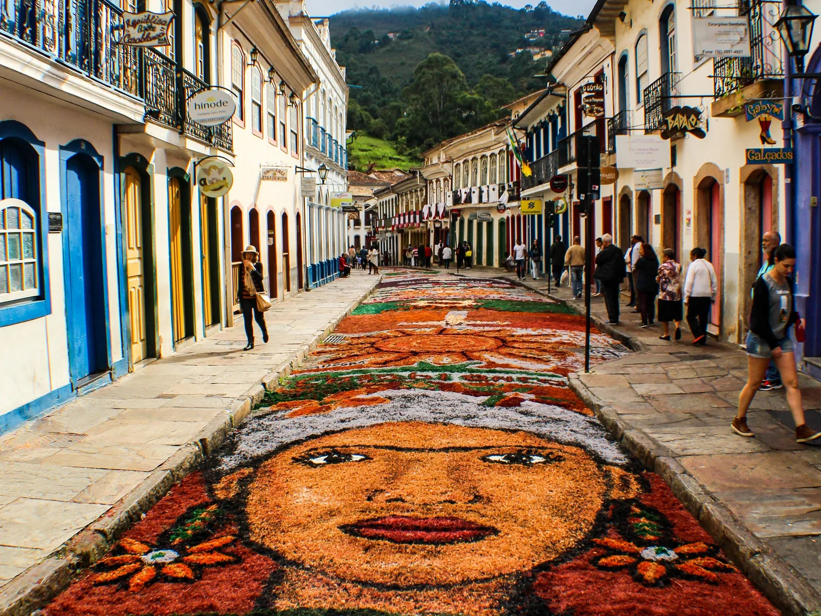 A devotional carpet laid down the middle of a cobbled Ouro Preto street for Holy Week, the face of Christ picked out in orange, red and green dyed sawdust, with people walking along the pavements either side between colonial houses