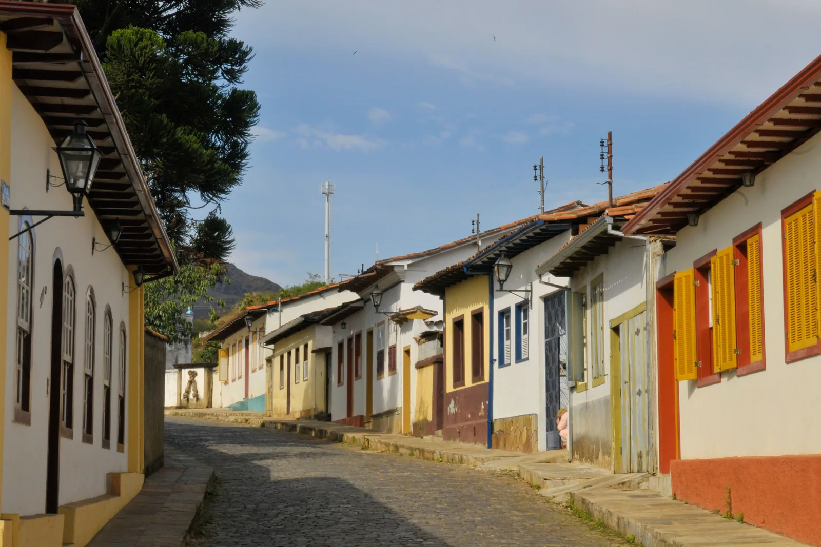 A quiet cobbled street in Mariana lined with single-story colonial houses painted white, yellow, orange and blue, with green hills at the end of it