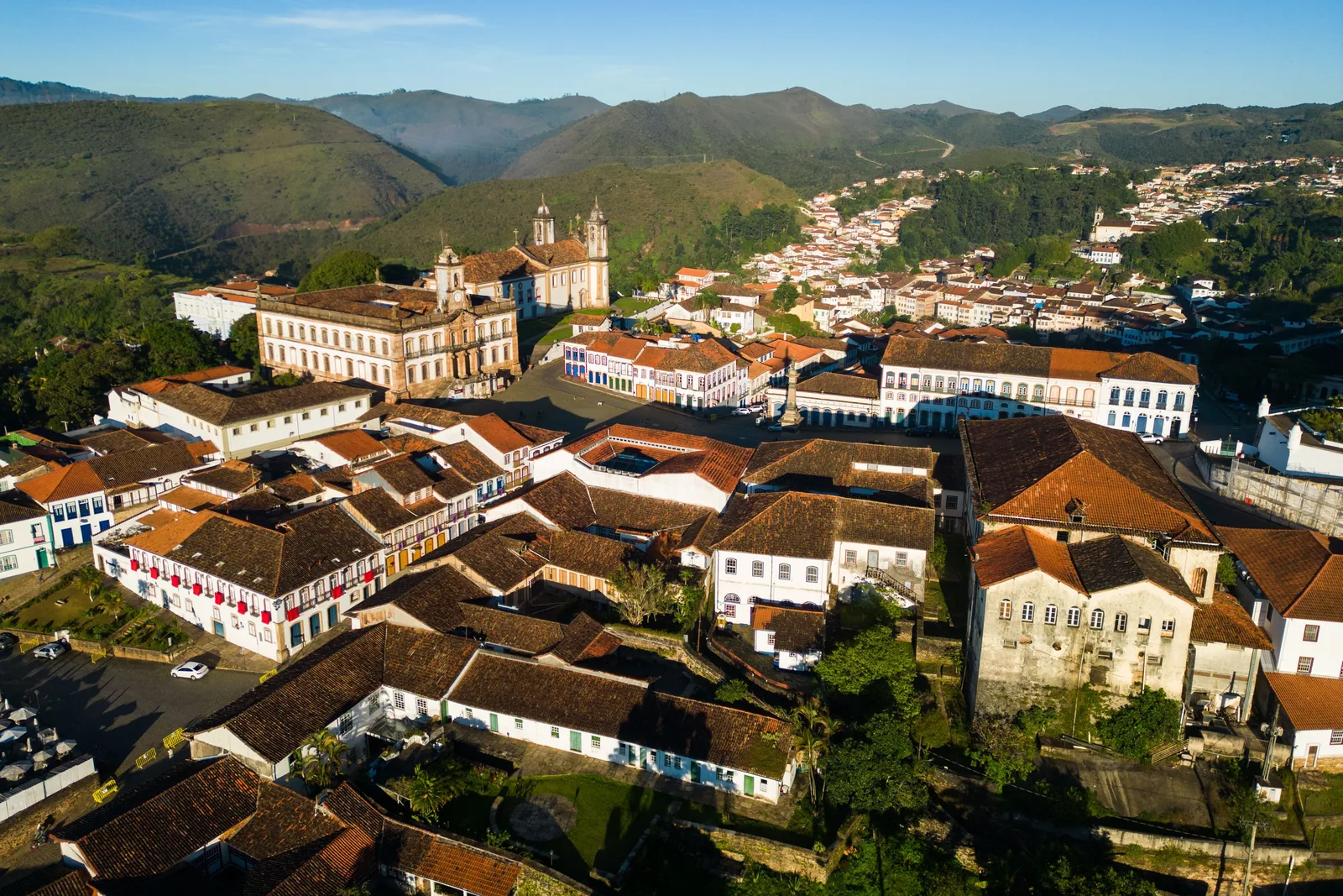 Ouro Preto from above: rows of red-tiled roofs and white walls running down the hillside, a church tower among them, and blue mountains along the skyline