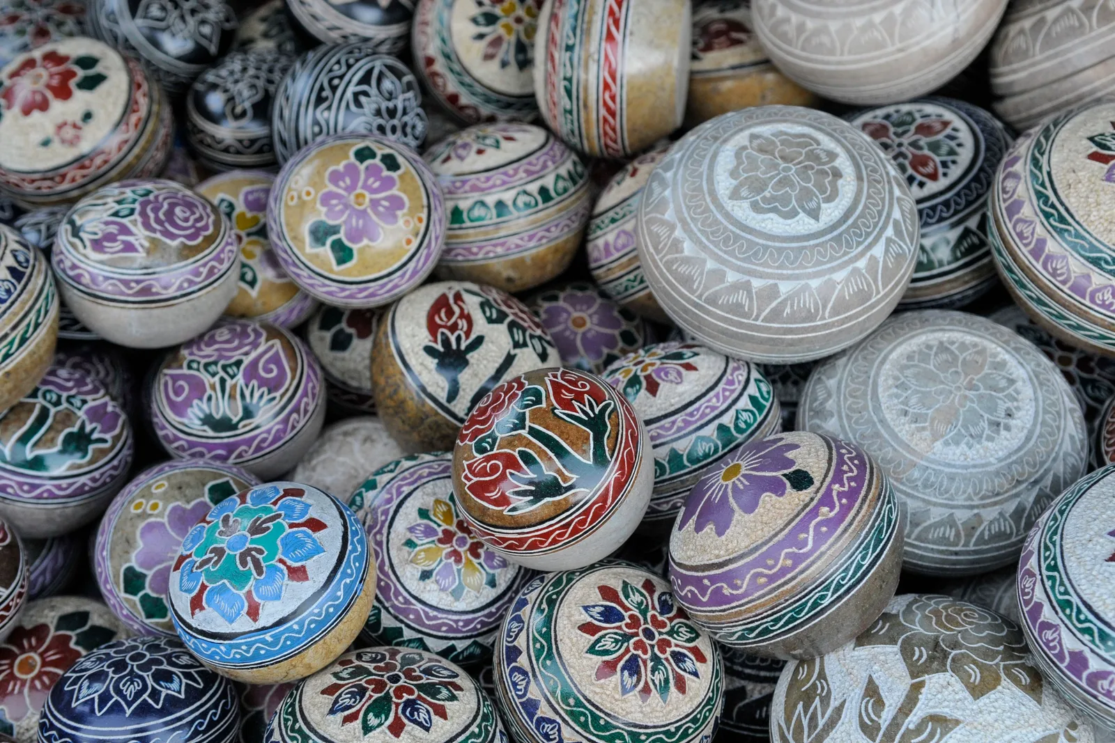 Dozens of round lidded soapstone pots stacked on a market stall, each carved and hand-painted in blue, green and pink floral patterns