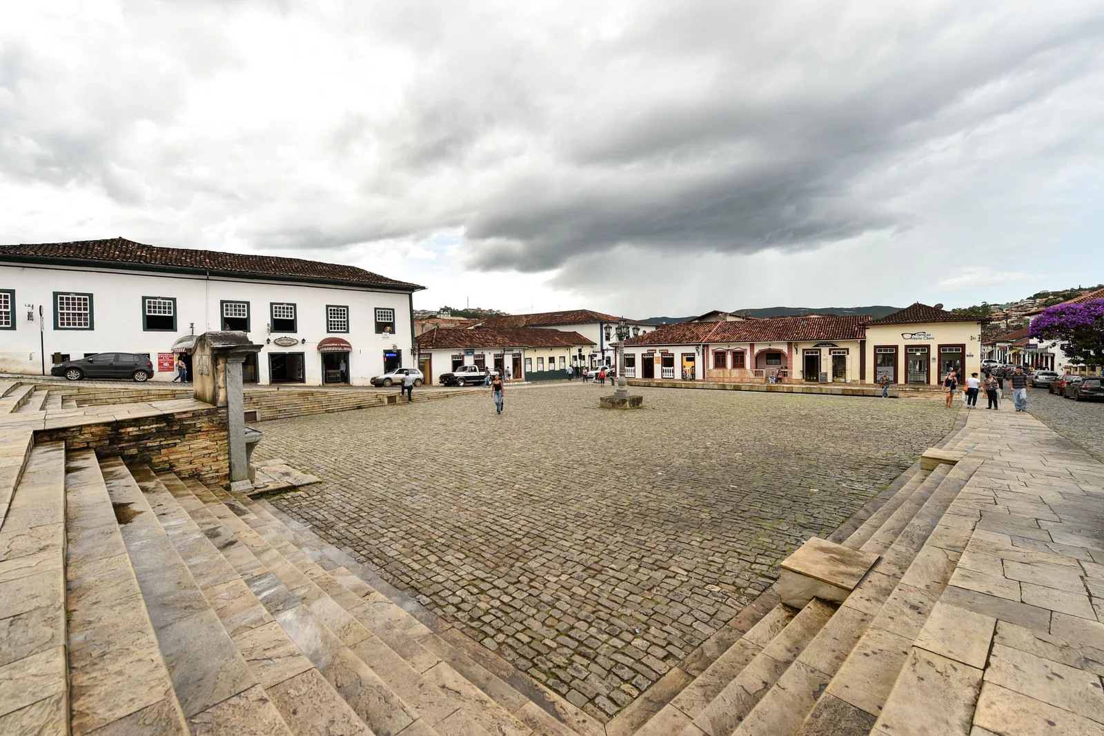The wide stone-paved Praça Claudio Manoel da Costa in Mariana on an overcast day, ringed by low white colonial buildings with arcaded fronts and almost empty of people