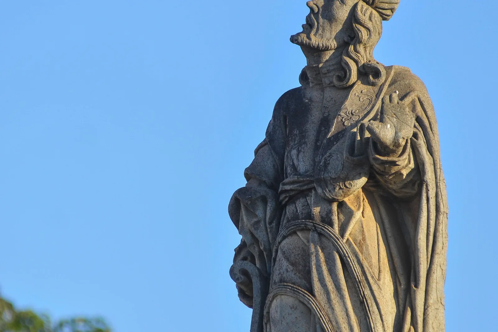 One of Aleijadinho’s soapstone prophets at Congonhas against a clear blue sky, a bearded figure in carved robes holding an open scroll, his name cut into the stone