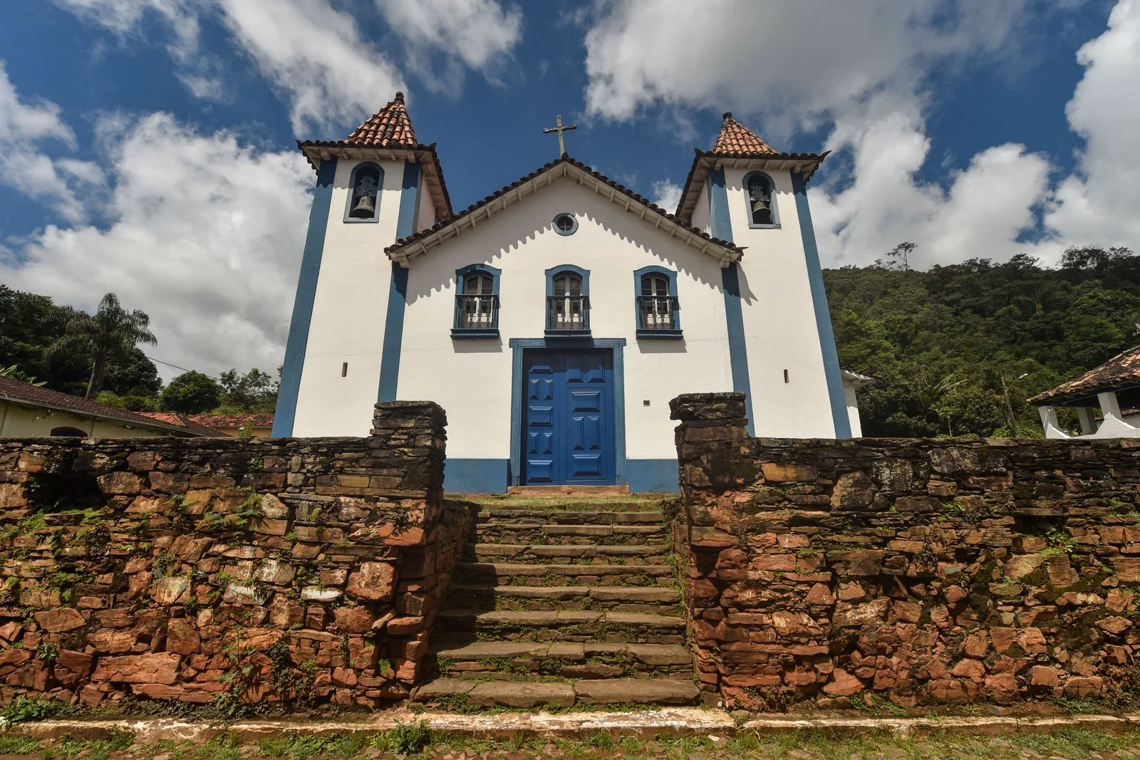 A small whitewashed chapel with a blue door and blue window frames at the top of a rough stone stairway in the rural district of São Bartolomeu, green hills behind it