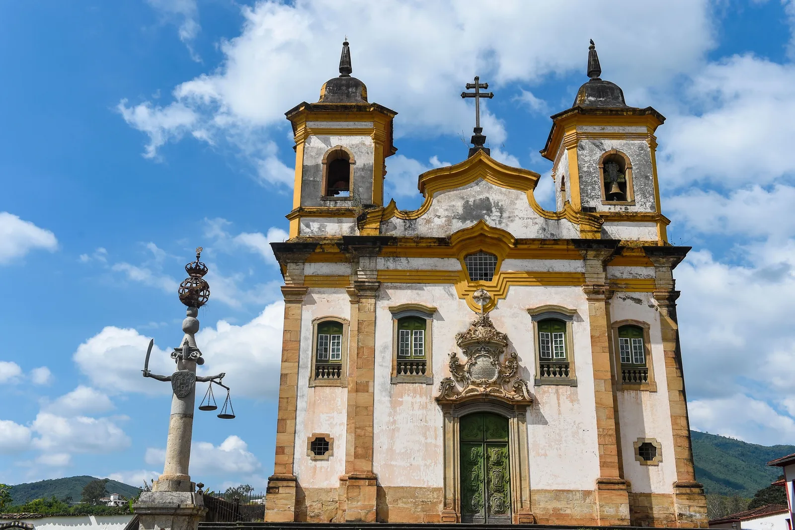 The Igreja de São Francisco de Assis on Mariana’s Praça Minas Gerais, twin bell towers and a carved stone portal against a blue sky, with the town’s stone pillory standing in front of it