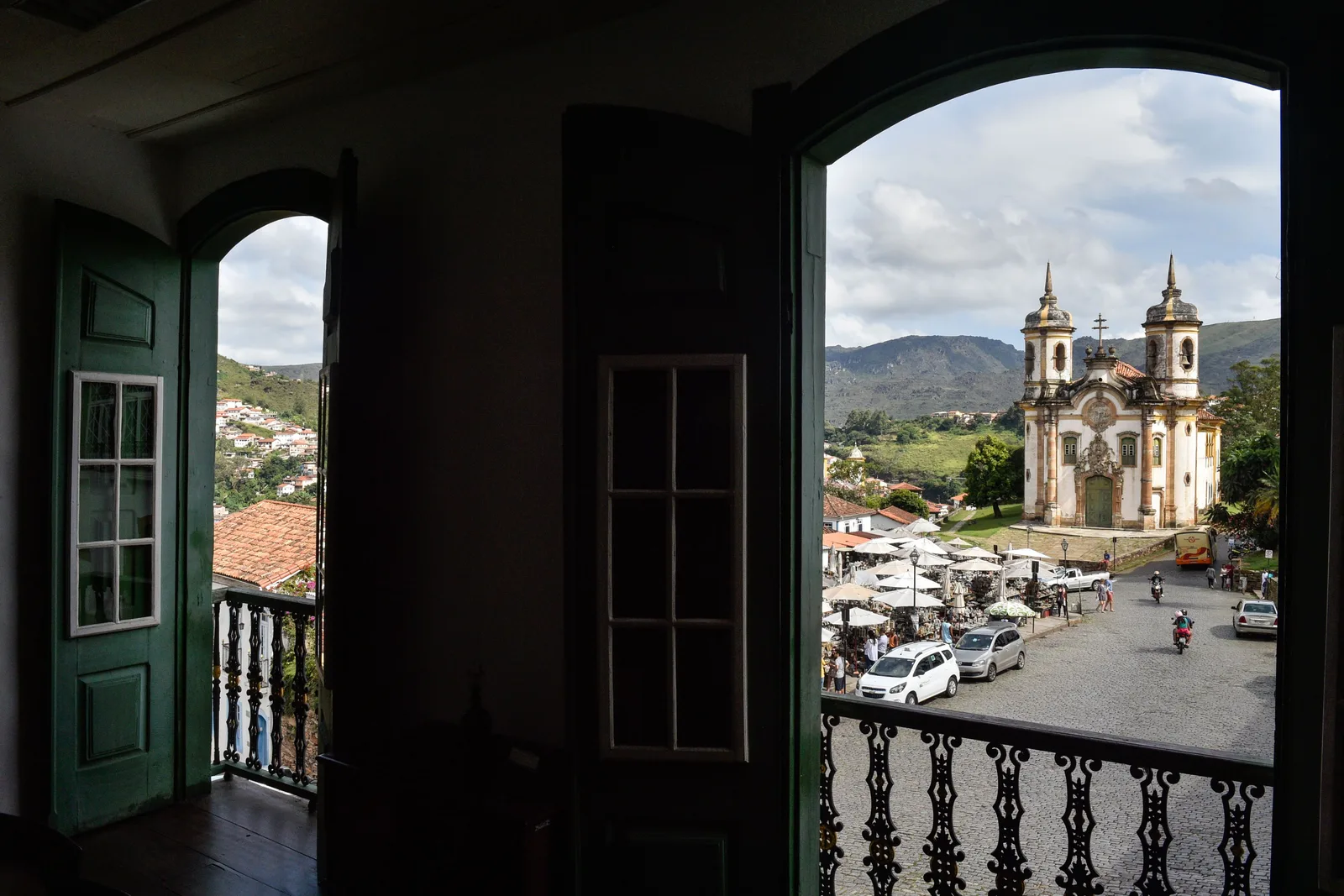The Igreja de São Francisco de Assis seen from a dark colonial interior through an open doorway and an iron balcony, the square below it full of parked cars and the hills beyond
