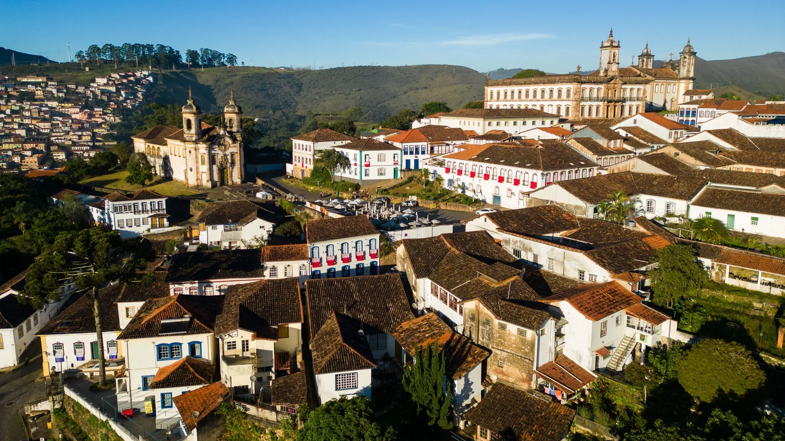 Ouro Preto from the air in early-morning light: white houses and red-tiled roofs packed down a steep hillside, a baroque church with twin domed bell towers standing on a green shoulder at the left, the long facade and clock tower of the Museu da Inconfidência ranged along the ridge opposite, and wooded hills behind