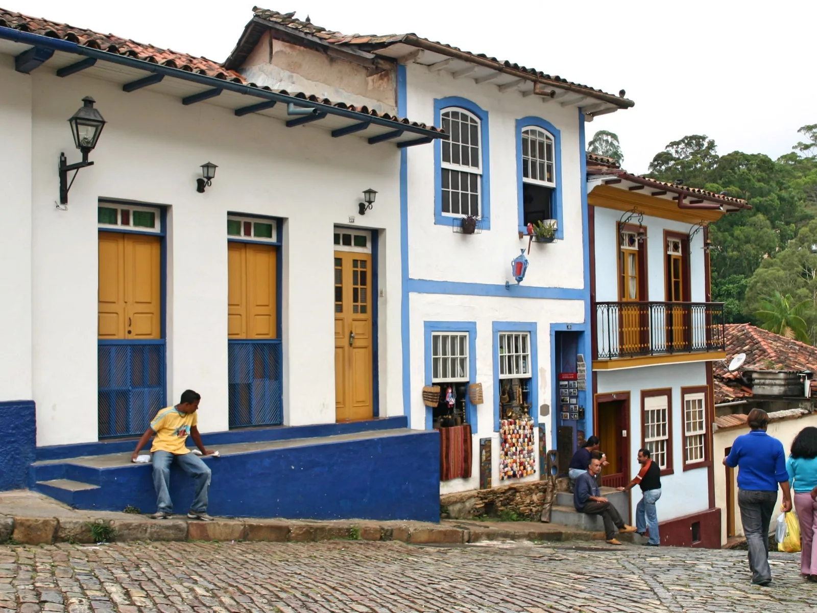 A steep cobbled street in Ouro Preto’s historic center lined with white colonial houses trimmed in blue, people sitting on the steps and walking uphill past shopfronts in strong midday sun