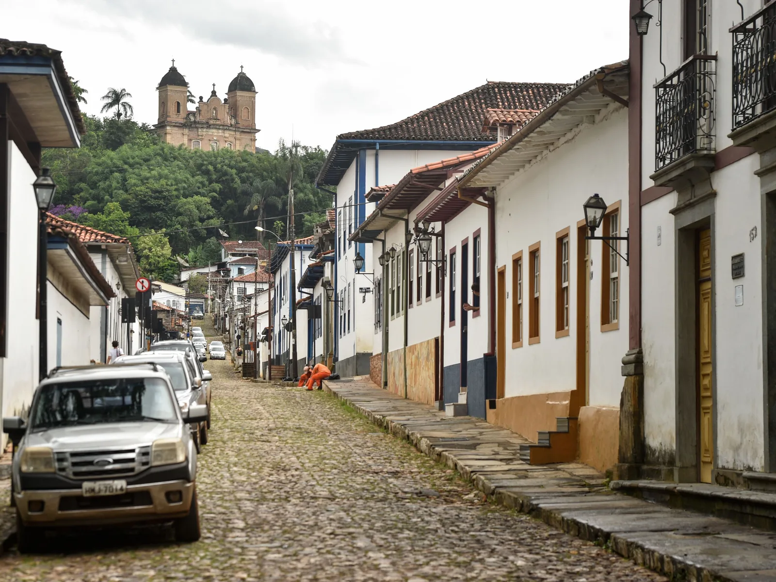 A cobbled street of white colonial houses in Mariana climbing gently toward the twin towers of the Basílica de São Pedro dos Clérigos on the hill above, cars parked along one side