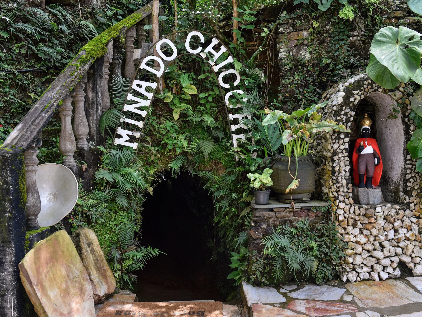 The entrance to the Mina do Chico Rei in Ouro Preto: a low dark tunnel mouth in a stone-faced wall overgrown with tropical plants, with the mine’s name spelled out in white letters on the rock above it