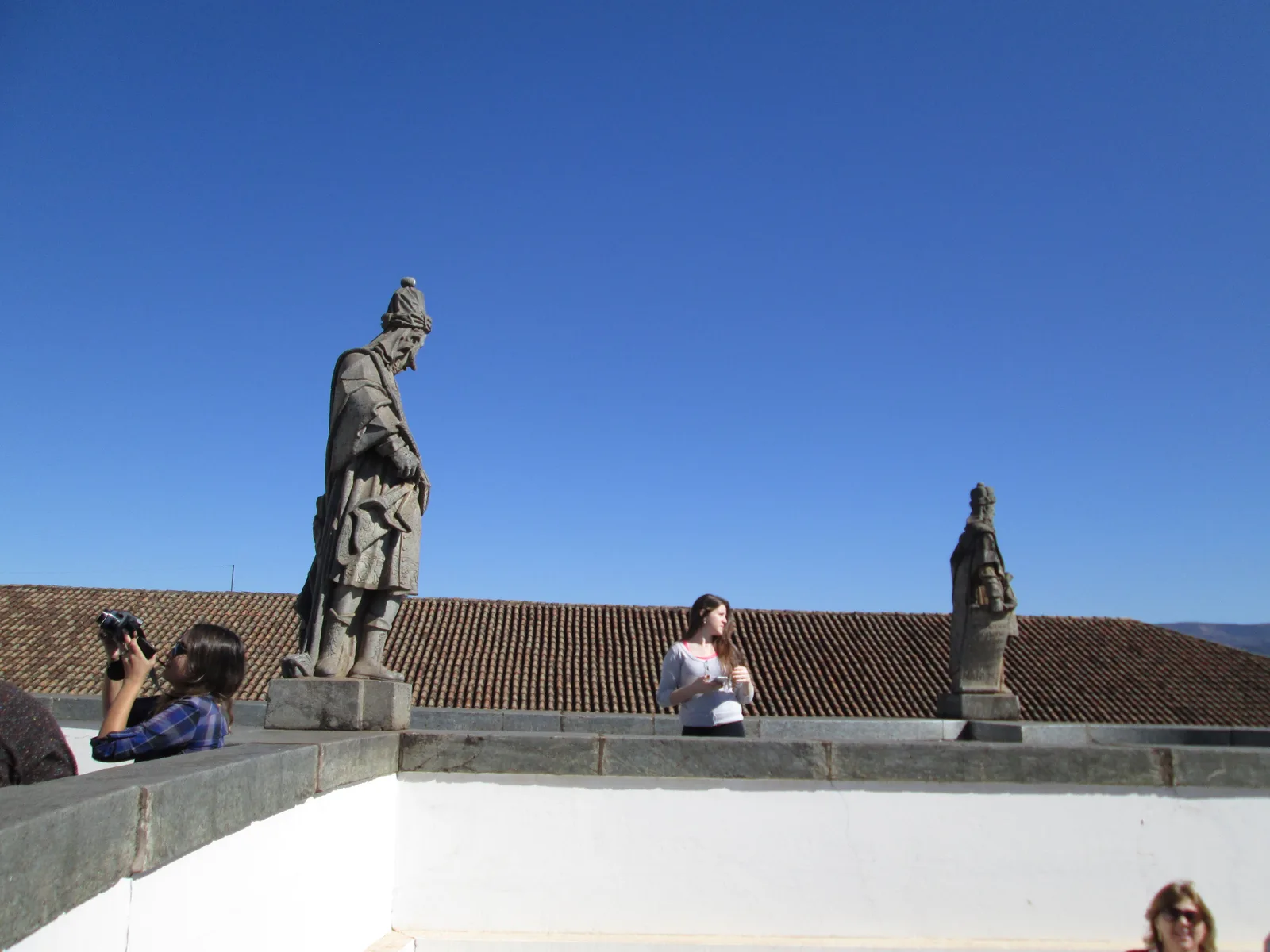 Two of Aleijadinho’s soapstone prophets standing on the terrace wall at Congonhas against a deep blue sky, visitors sitting and taking photographs on the low wall between them