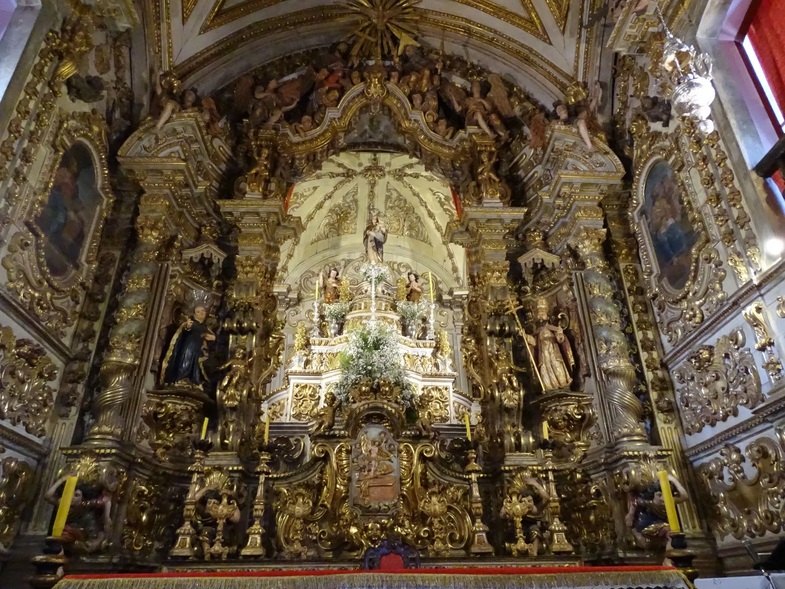 The high altar of the Basílica de Nossa Senhora do Pilar in Ouro Preto: tiers of gilded baroque carving rising into the vault, gold columns and scrollwork on every surface, painted saints standing in niches either side, and a small lit image at the center