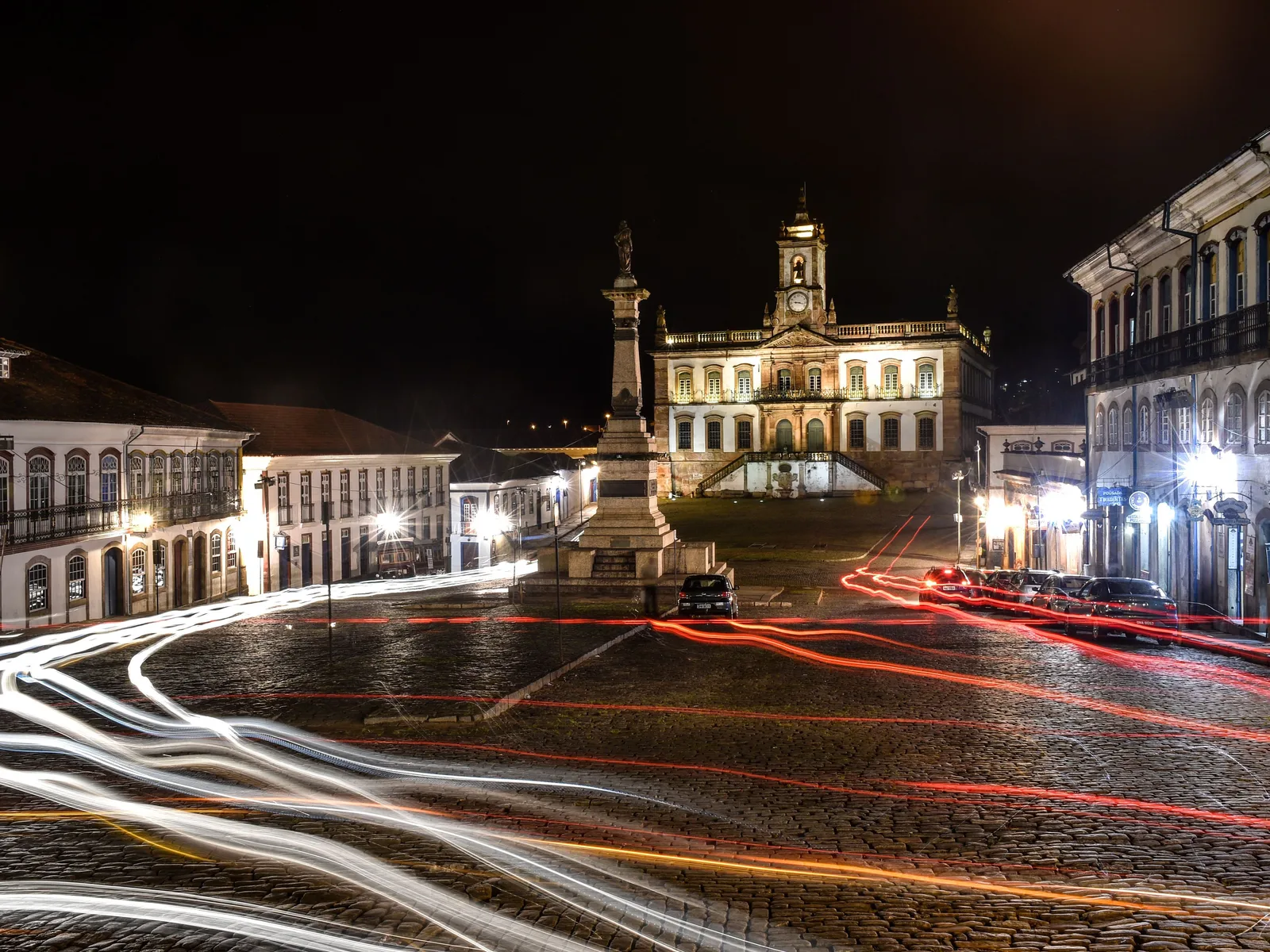 Praça Tiradentes at night in a long exposure: the floodlit Museu da Inconfidência standing at the head of the square with the lit Tiradentes column in front of it, and red and white streaks of car light curving across the wet cobbles in the foreground
