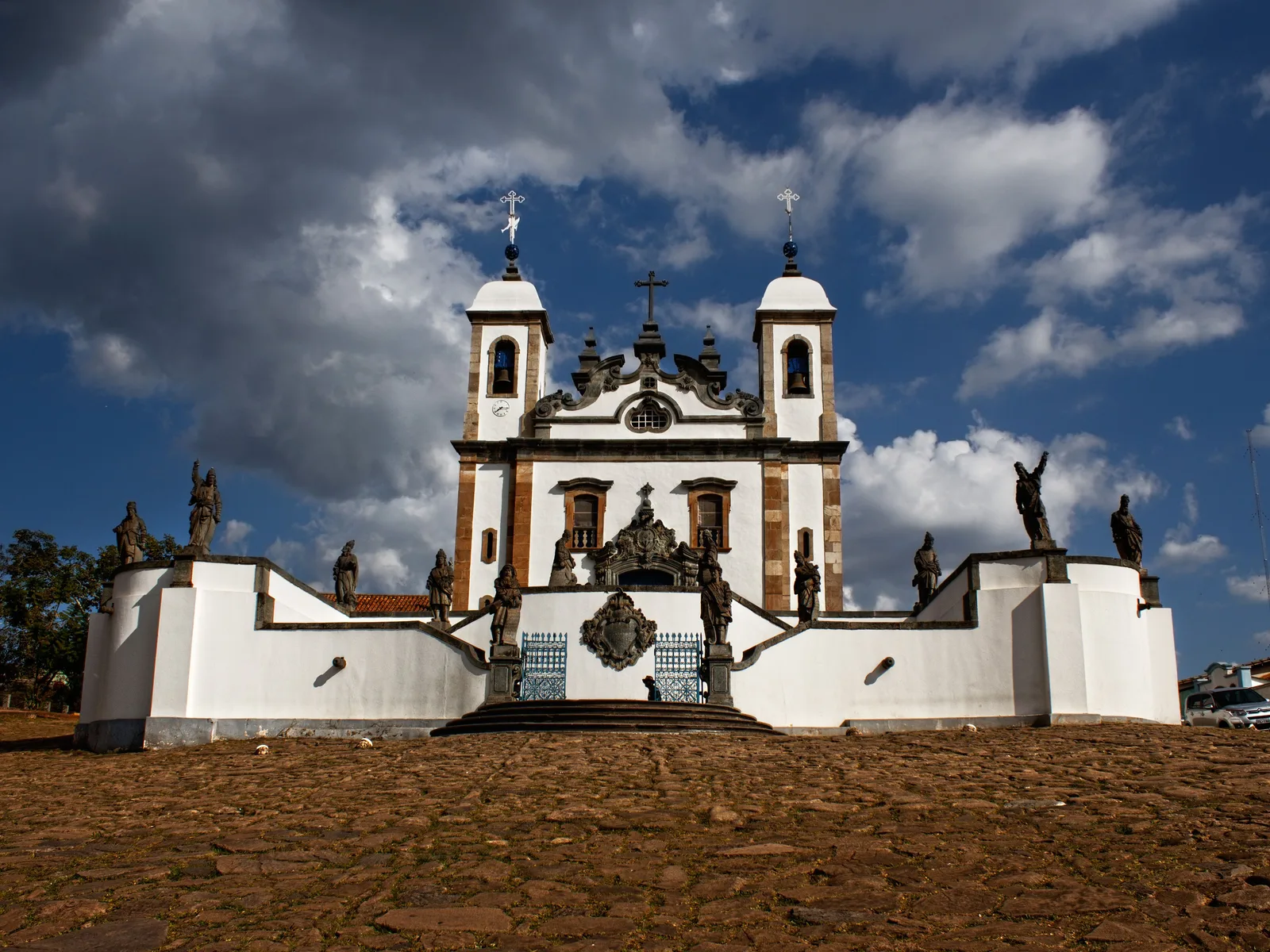 The Santuário do Bom Jesus de Matosinhos at Congonhas under heavy cloud: a white baroque church with two bell towers behind a curving terrace wall, Aleijadinho’s soapstone prophets standing along the balustrade and either side of the double stone stairway