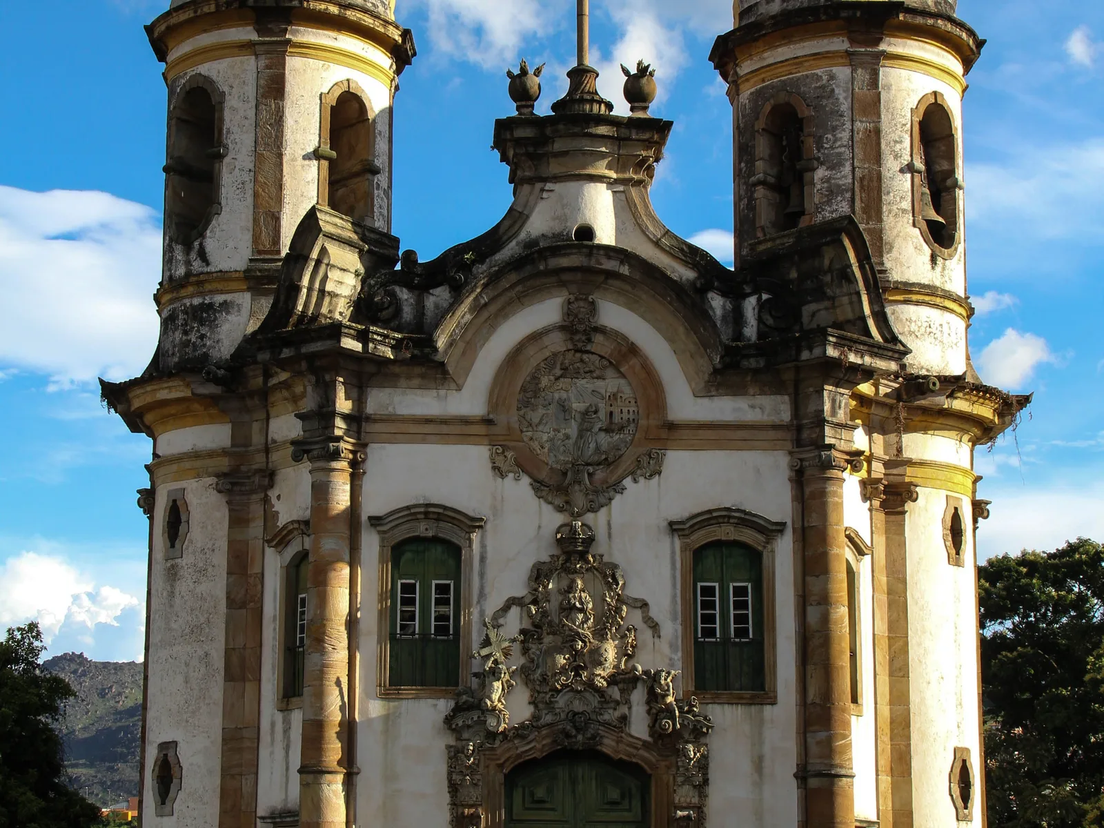 The upper facade of the Igreja de São Francisco de Assis in Ouro Preto against a blue sky: two cylindrical bell towers with domed caps flanking a scrolled white pediment, and Aleijadinho’s deeply carved soapstone medallion set above the doorway between green-shuttered windows