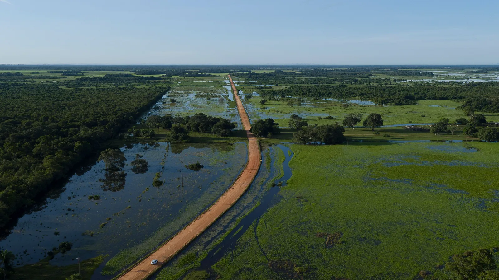 Aerial view of a raised dirt causeway curving away across a flooded green plain toward a flat horizon, a long dark channel of standing water beside it and a single small white vehicle on the road