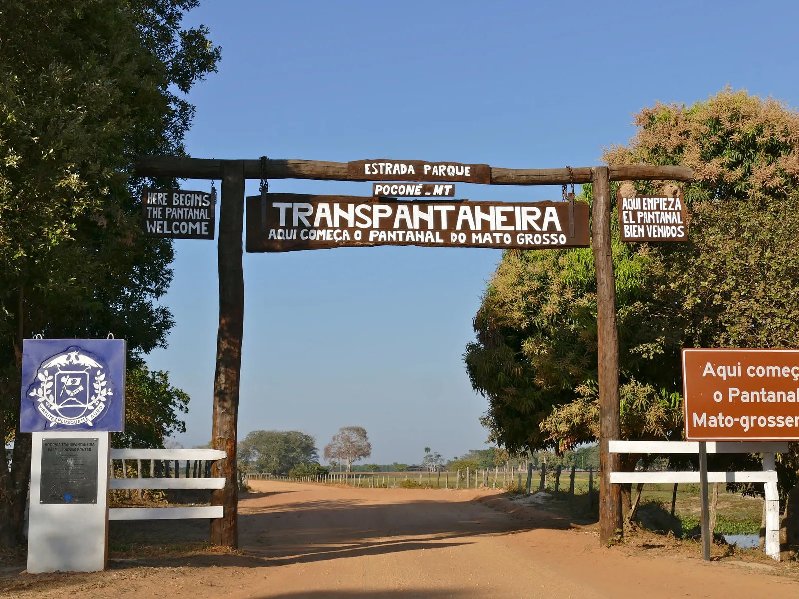 A timber arch over a red dirt road lettered “Estrada Parque, Poconé - MT, Transpantaneira — aqui começa o Pantanal do Mato Grosso”, with smaller welcome boards in English and Spanish on its posts, the Mato Grosso coat of arms on a banner at the left and white rails either side of the road