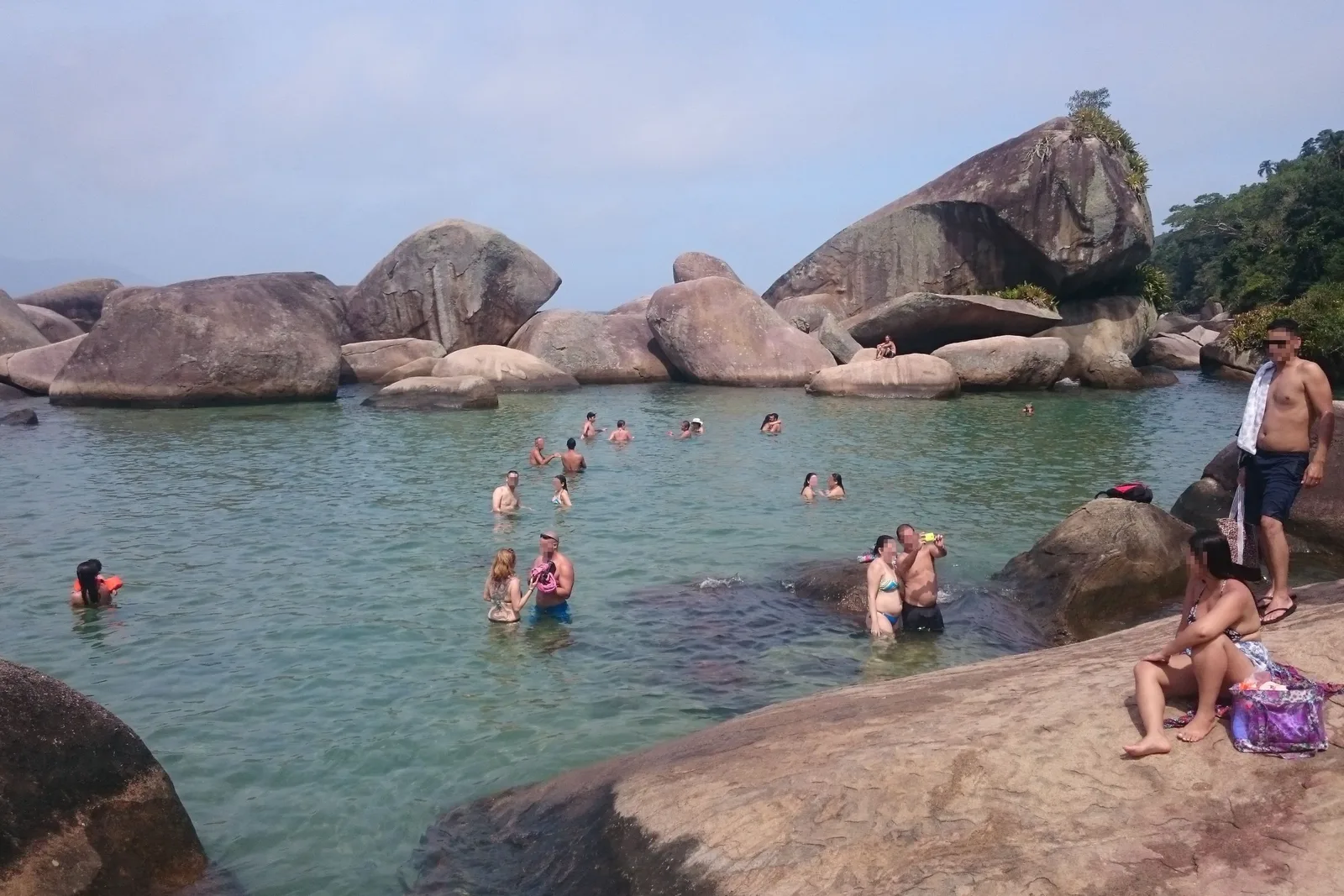 The Cachadaço natural pool at Trindade: turquoise water held inside a ring of dark boulders with swimmers in it, ocean swell breaking on the rocks just beyond and forest rising behind