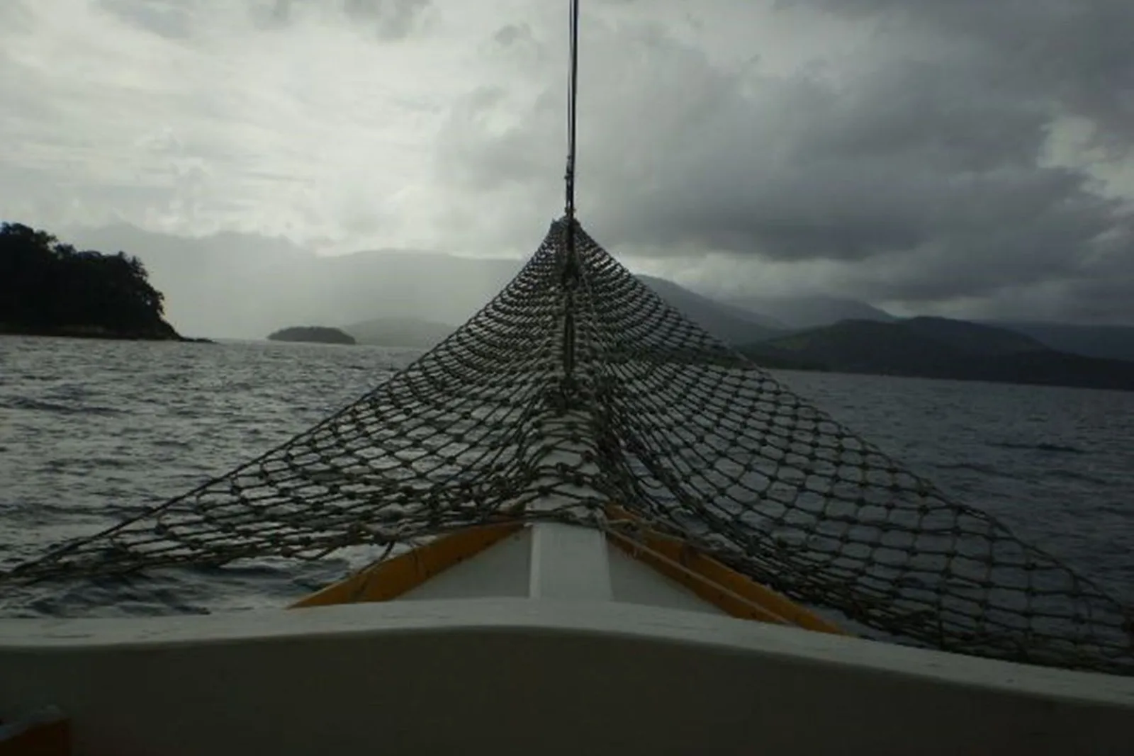 The bow of a small boat crossing Paraty bay under a heavy sky, forested islands low on the horizon beside it
