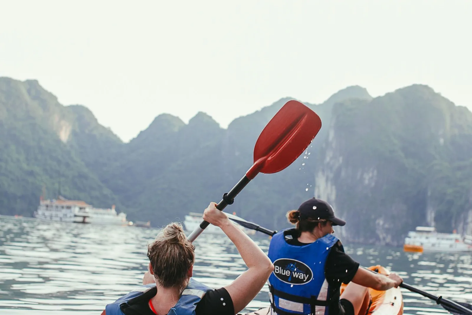 Two people kayaking in tandem on flat water below steep forested slopes