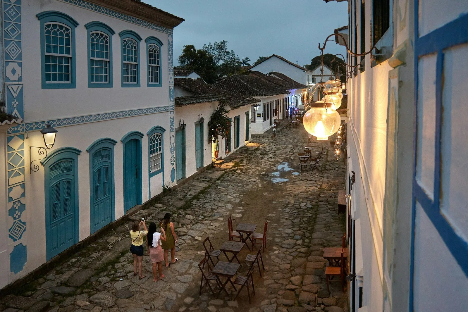 Tables and chairs set out on a Paraty cobbled street at dusk, lamps lit along the whitewashed colonial facades and people walking between them