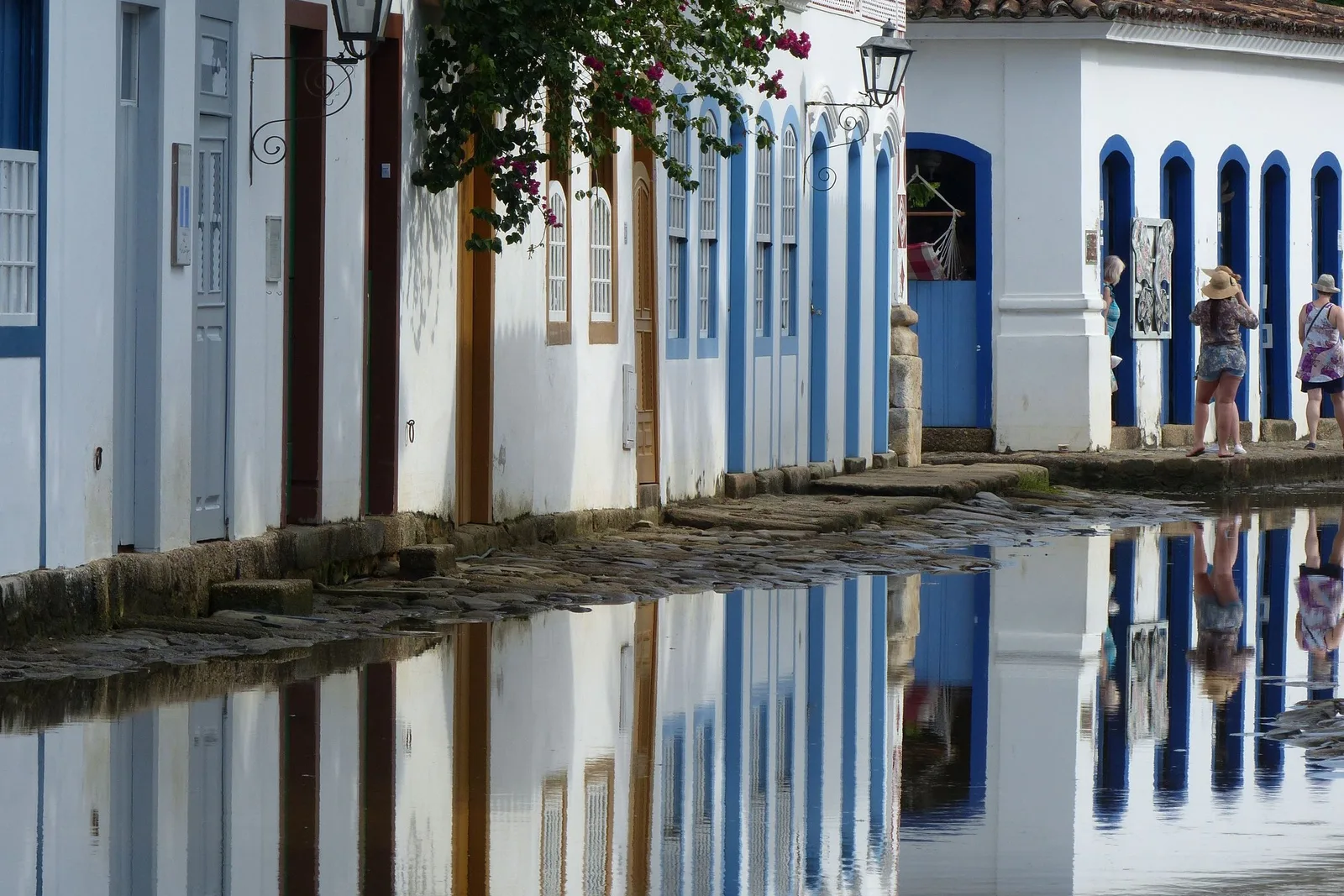 People walking barefoot through shallow seawater covering a Paraty street at high tide, the colonial houses and a lit doorway mirrored perfectly in the still water at dusk