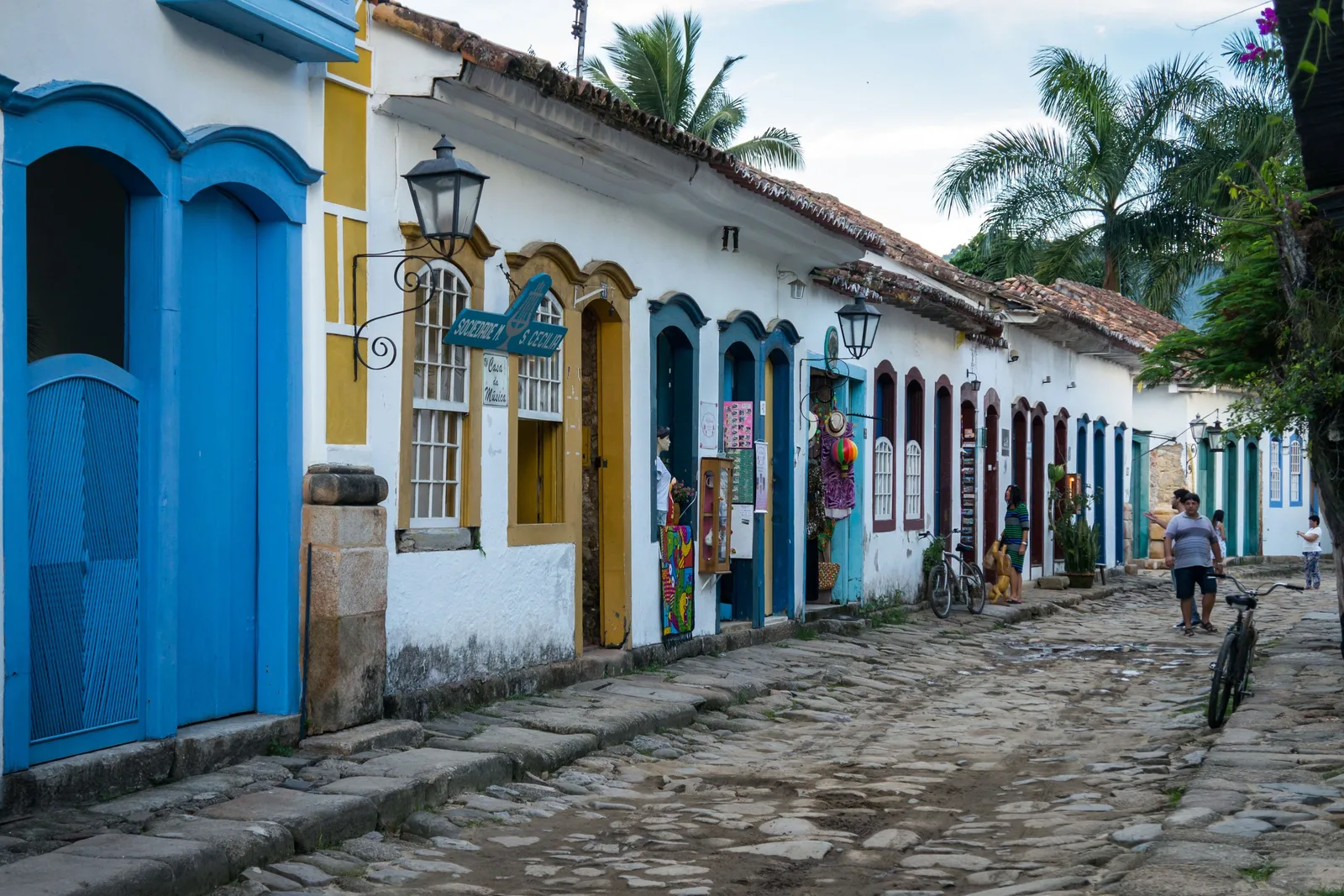 A narrow Paraty street in low late-afternoon light, whitewashed houses with blue and ochre window frames on both sides and the rough stone paving running away toward a church