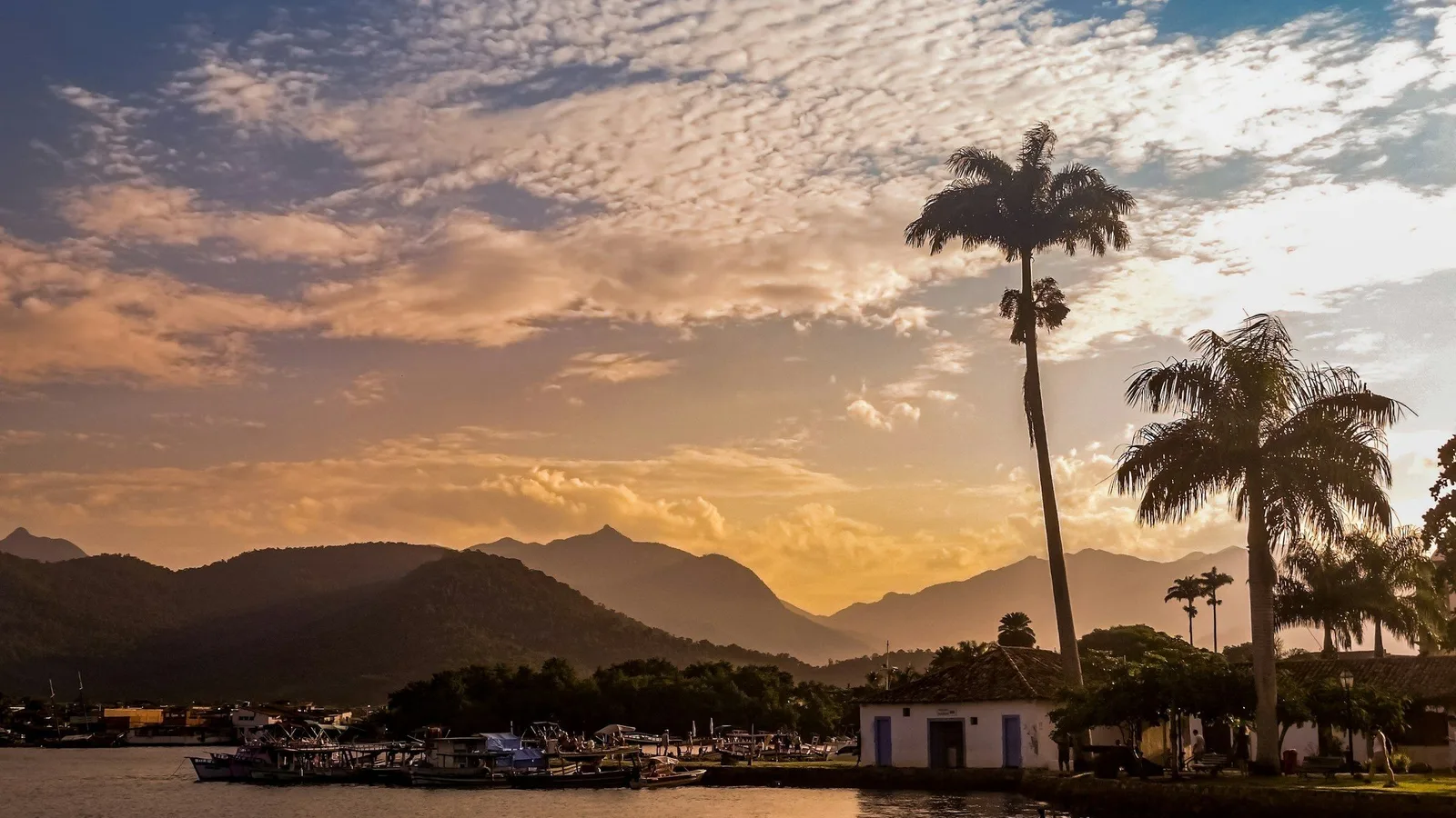 Paraty bay at sunset from the water, wooden schooners silhouetted at anchor and the dark ridges of the Serra do Mar layered behind them under a burning sky