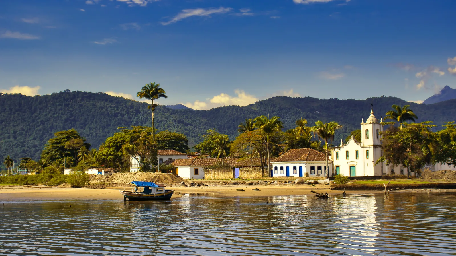 Aerial view of Paraty at golden hour: the white-and-color grid of the colonial center with the Matriz church tower at its heart, the Perequê-Açu river mouth on one side, and the bay and its wooded islands behind, with the wall of the Serra do Mar rising steeply behind the town
