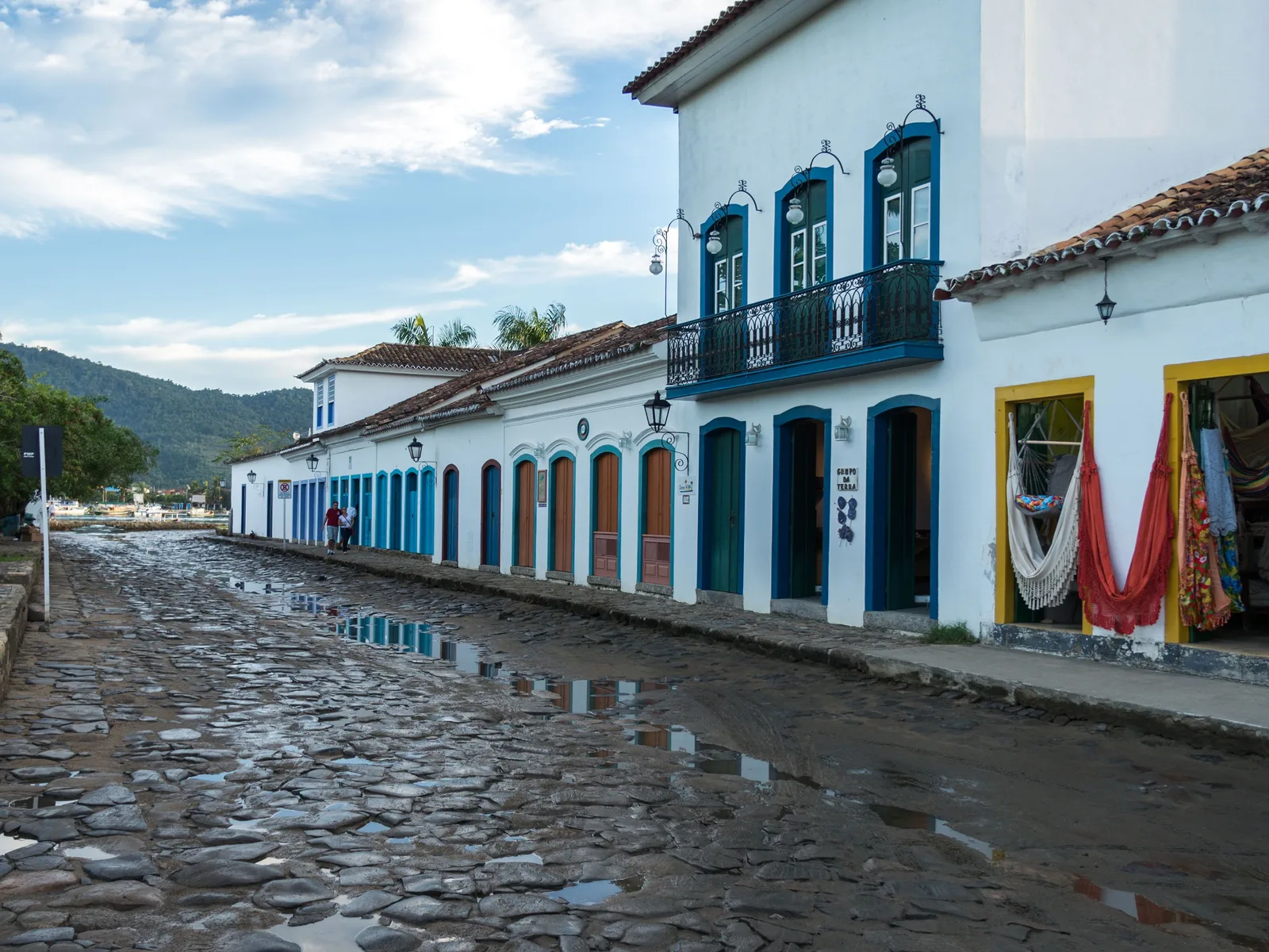 Paraty’s main square at dusk with the floodlit neoclassical Matriz church, a few people crossing the rough stone paving and warm light in the doorways of the houses around it