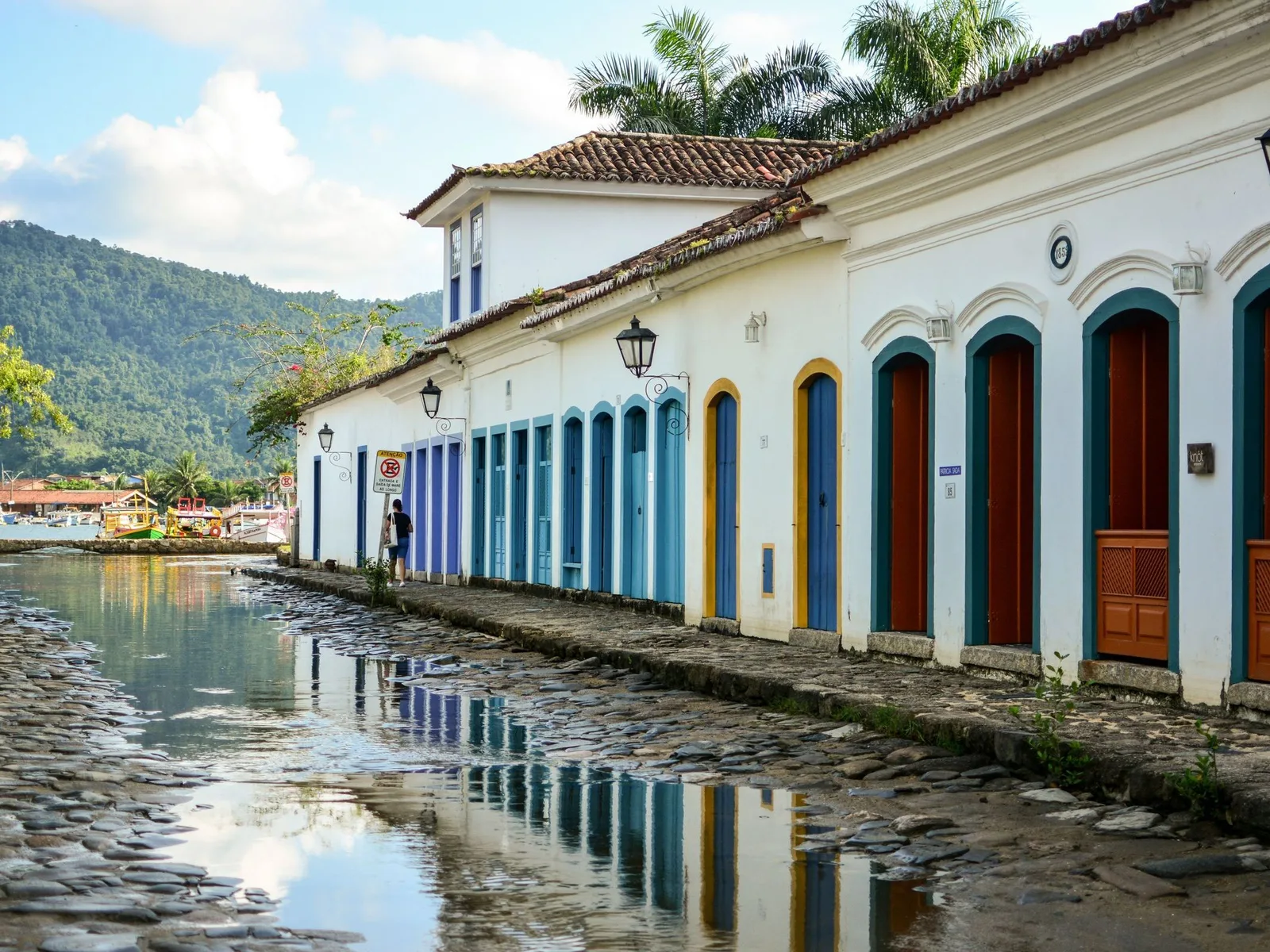 A Paraty street at high tide: seawater standing ankle-deep over the rough stone paving, whitewashed colonial houses with brightly painted door frames reflected upside down in it, and a church tower at the end of the street