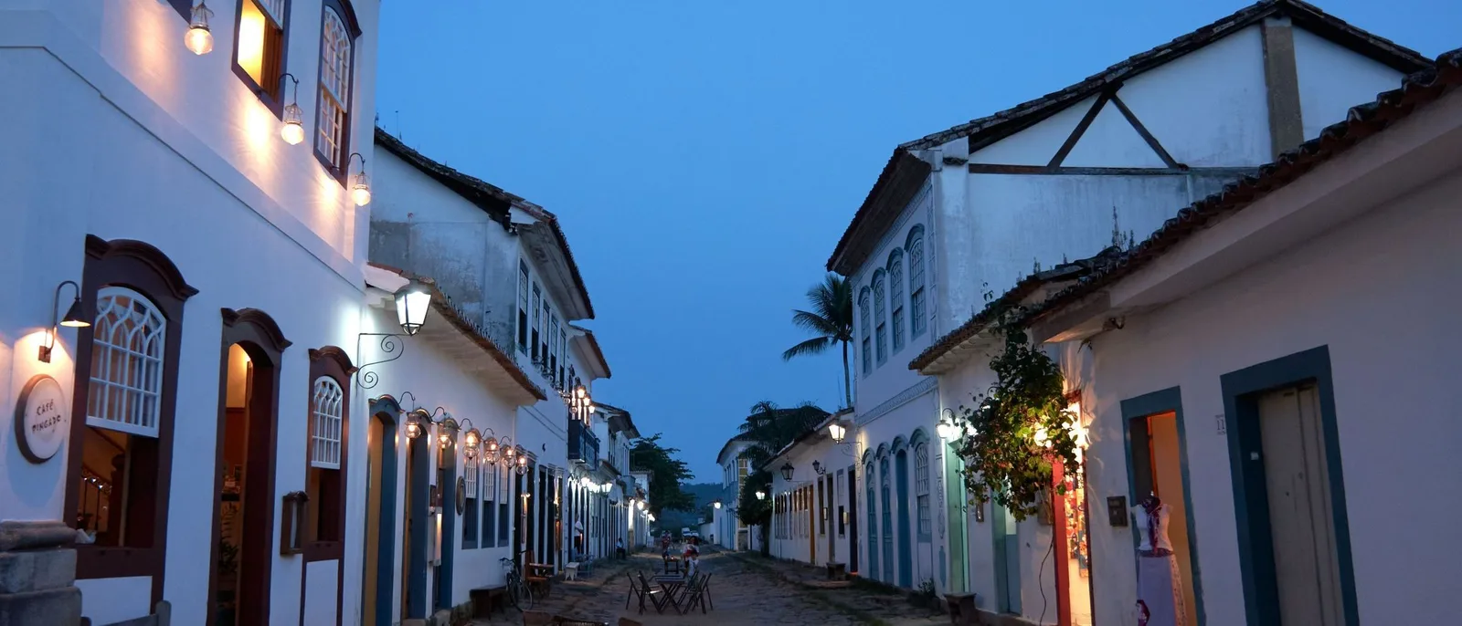 A cobbled Paraty street at dusk, whitewashed houses with brightly painted doors and window frames on both sides, warm light spilling from open doorways and the church tower closing the far end