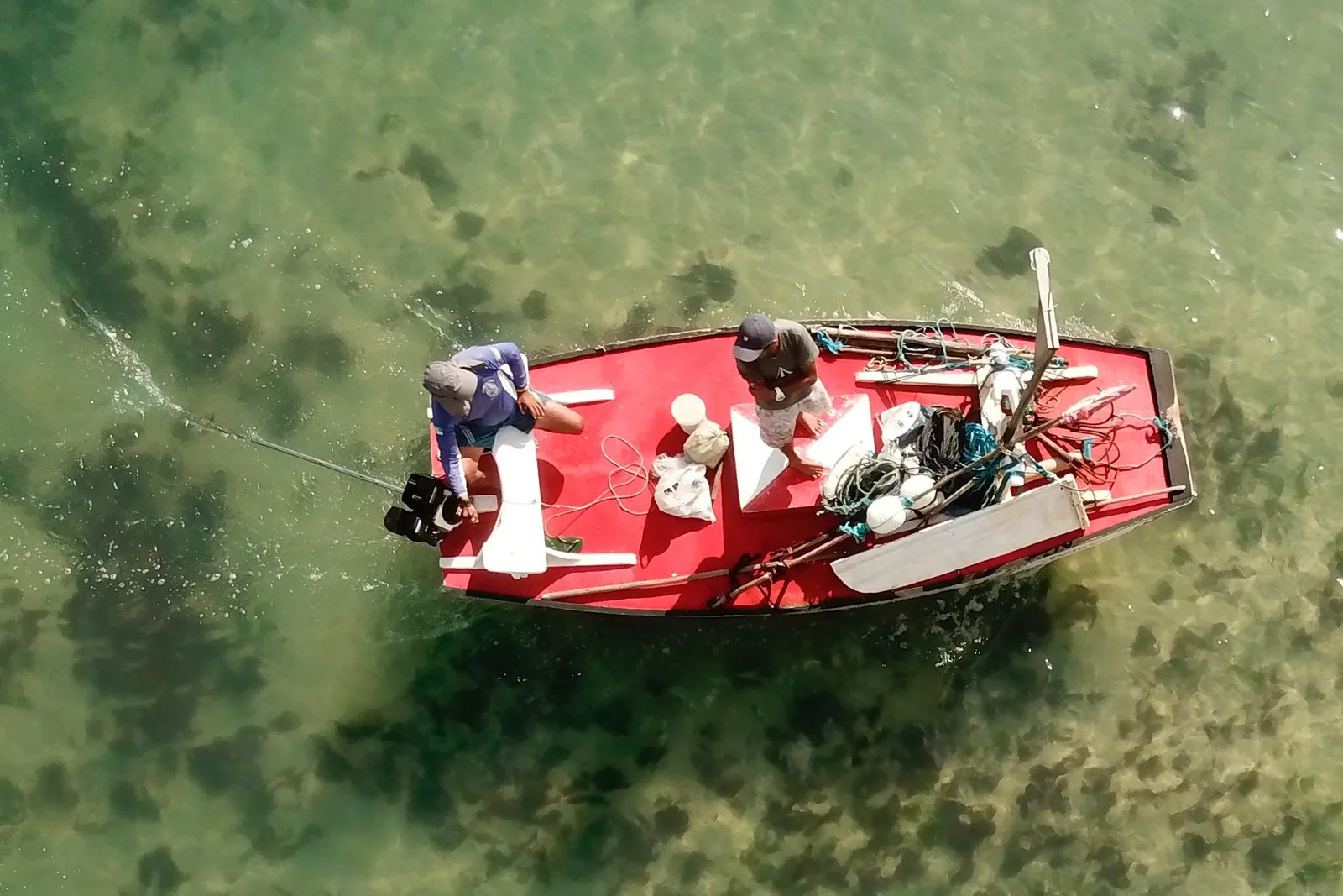 A small red wooden fishing boat photographed from directly above, two men aboard among nets, floats and rope, on clear green water over sand and dark weed