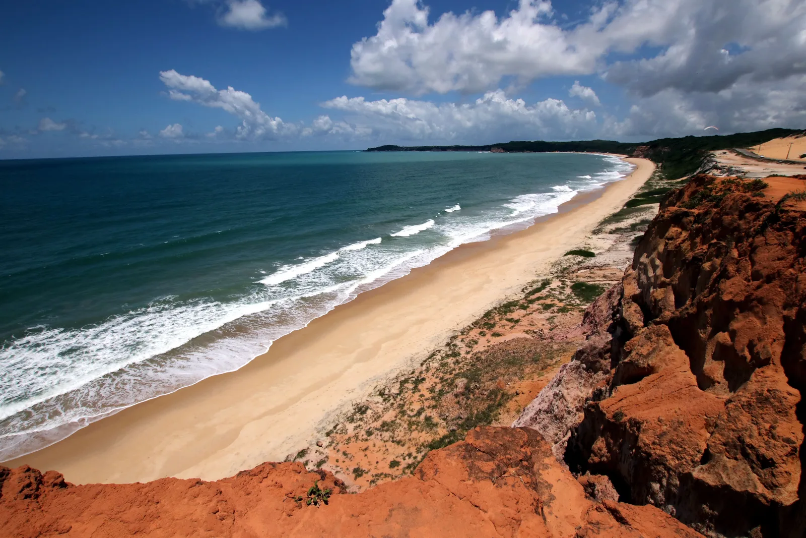 A long curving beach seen from a red clifftop at Cacimbinhas, lines of surf running up the pale sand, green scrub along the cliff edge and dunes in the distance