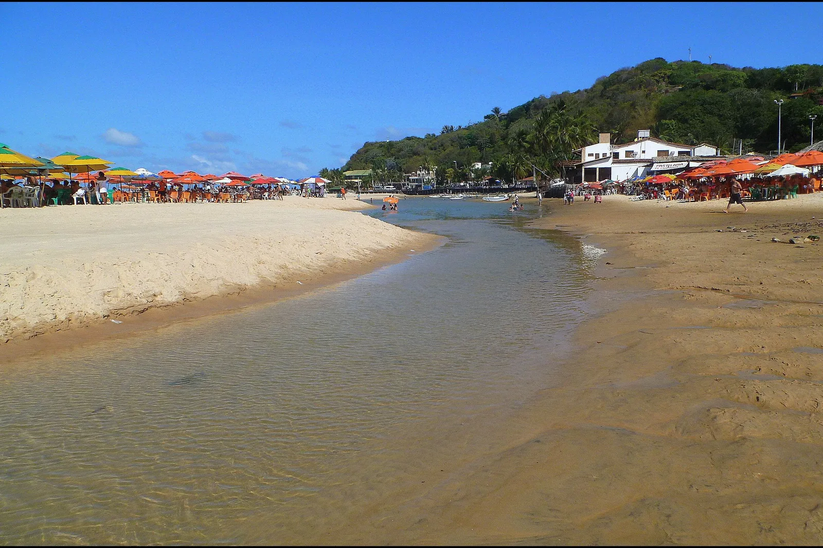 The beach in front of Pipa at low tide, a shallow channel of water running across wet sand with parasols and buildings along the shore and a green headland beyond