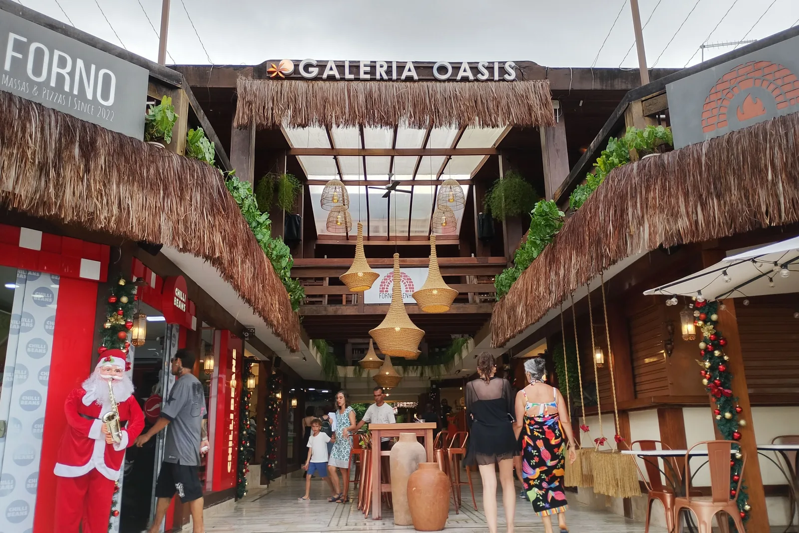 A covered shopping arcade in Pipa lined with small shopfronts under thatched eaves, woven lamps hanging overhead, Christmas decorations and a Santa figure at the entrance, and people walking through it