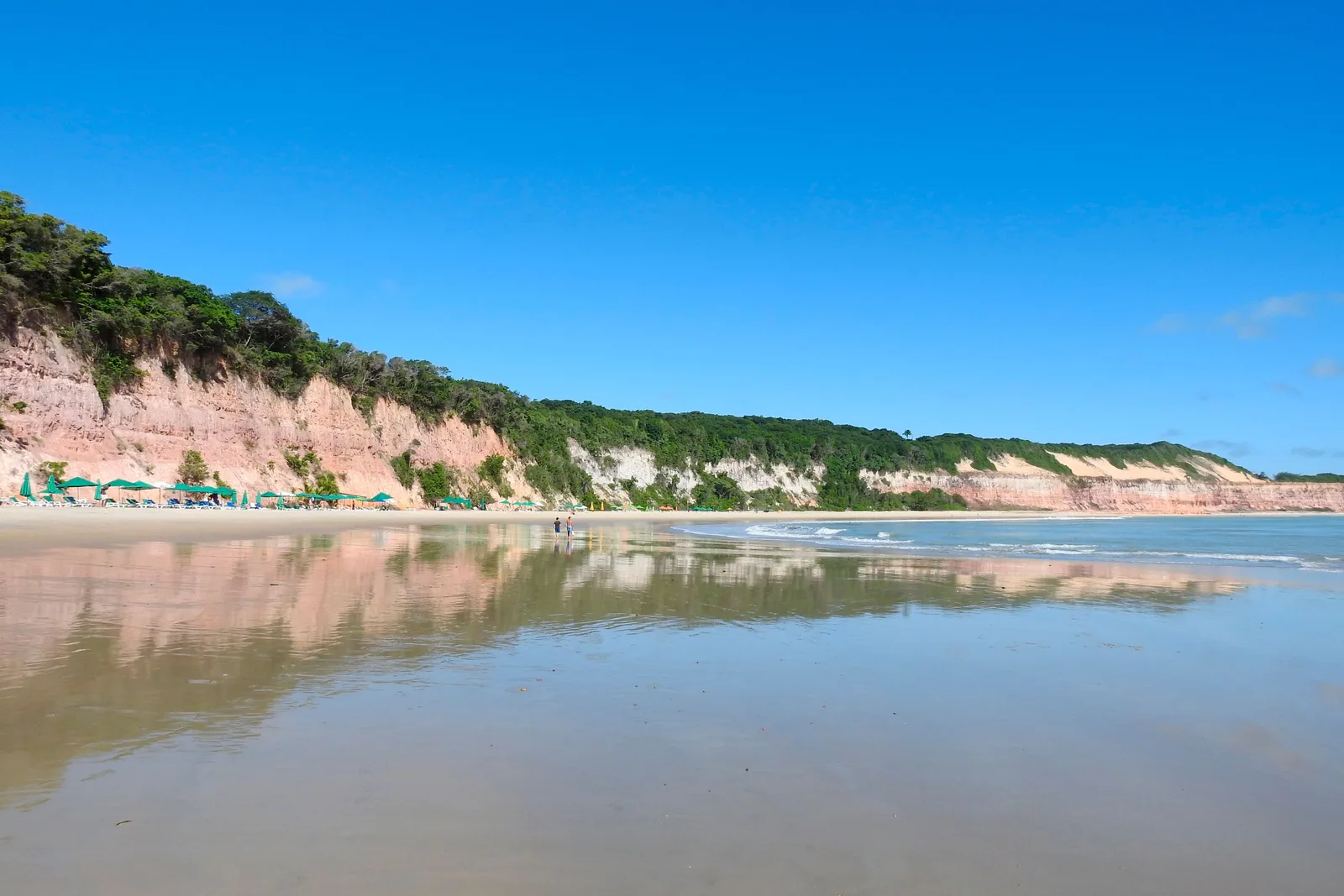 Wet sand in Baía dos Golfinhos reflecting the pink and white cliffs above it, a row of green parasols at the foot of the cliff and two people at the water’s edge